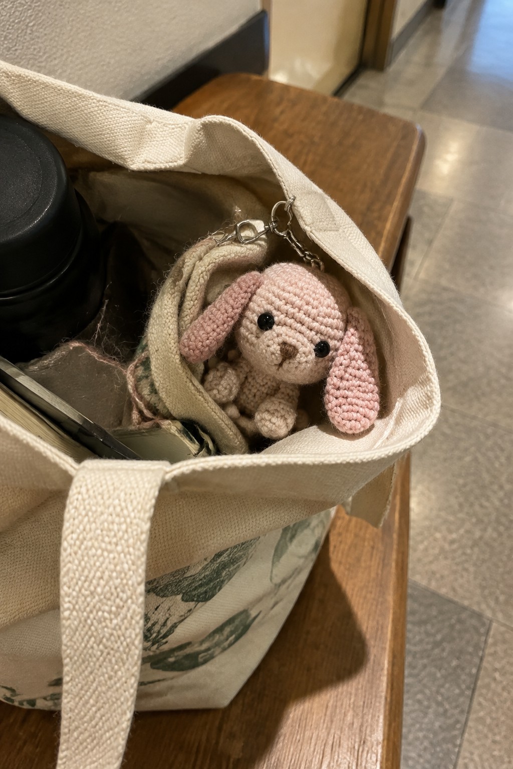 Pink crocheted bunny keychain on canvas tote bag with black thermos on bench