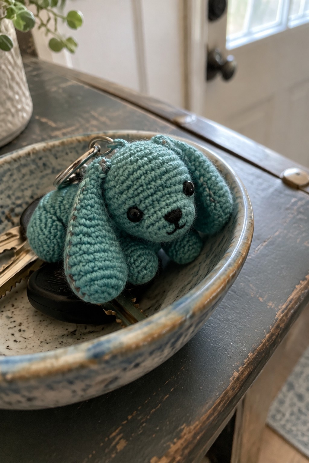 A small turquoise crocheted dog keychain with floppy ears sits in a blue ceramic bowl on a wooden table.