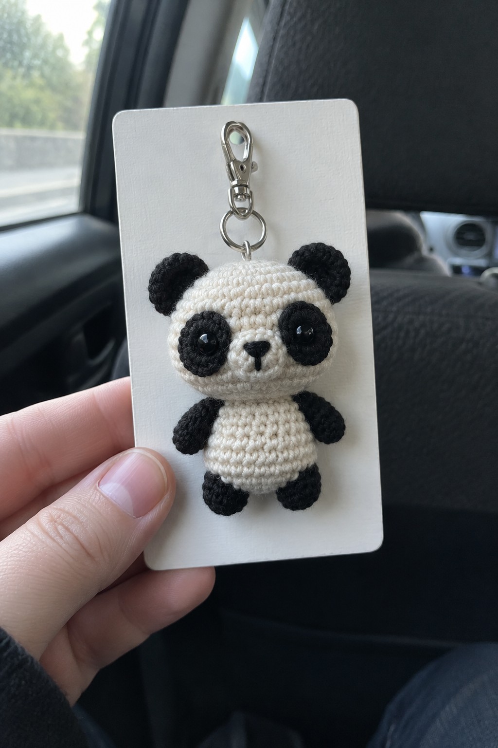 Hand holding white card with black-and-white crocheted panda keychain in car
