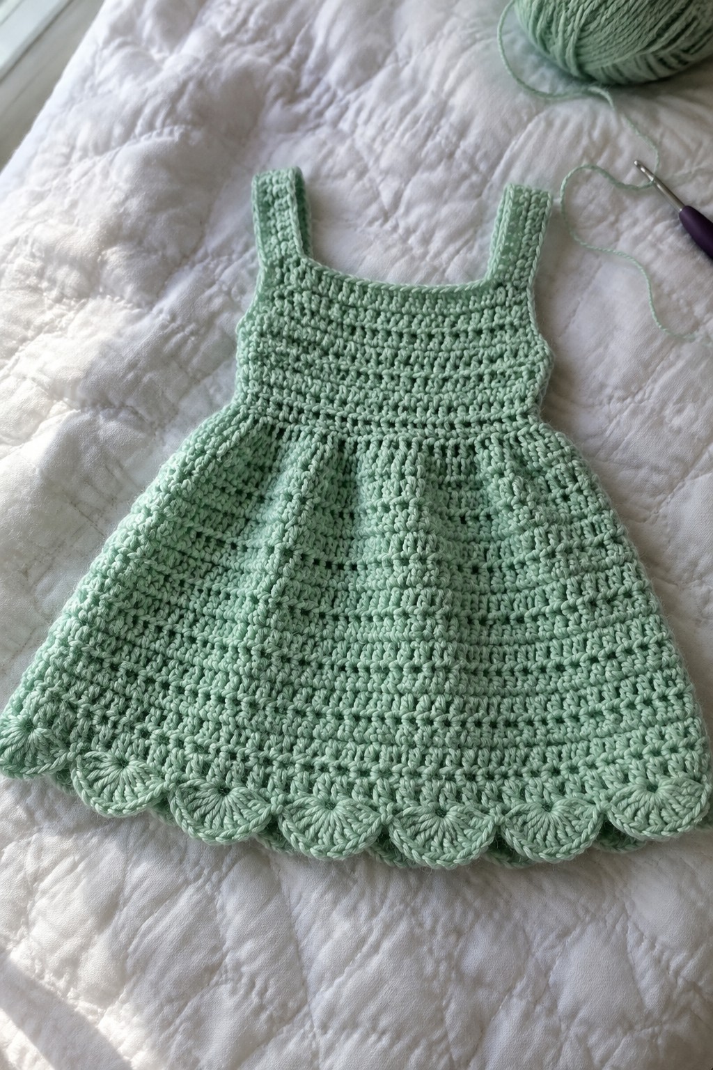 Mint green crocheted dress with scalloped hem on white quilt beside yarn and hook
