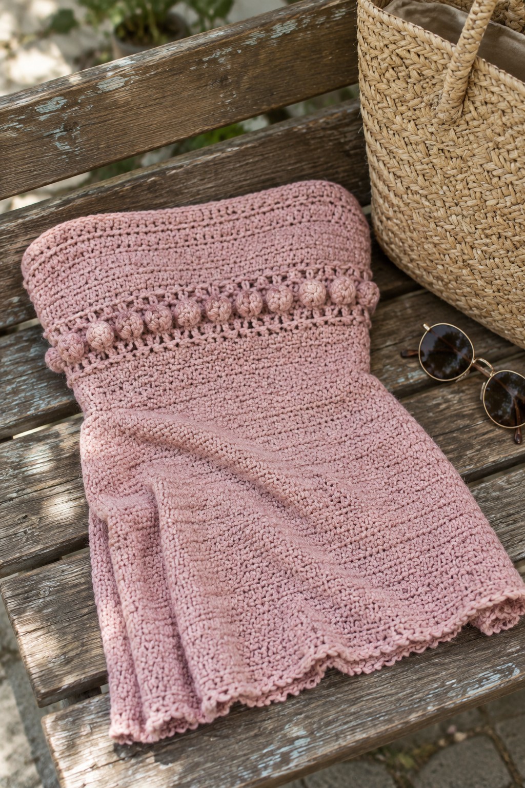 Pink knitted strapless dress with pom-pom trim on wooden bench beside straw bag and sunglasses