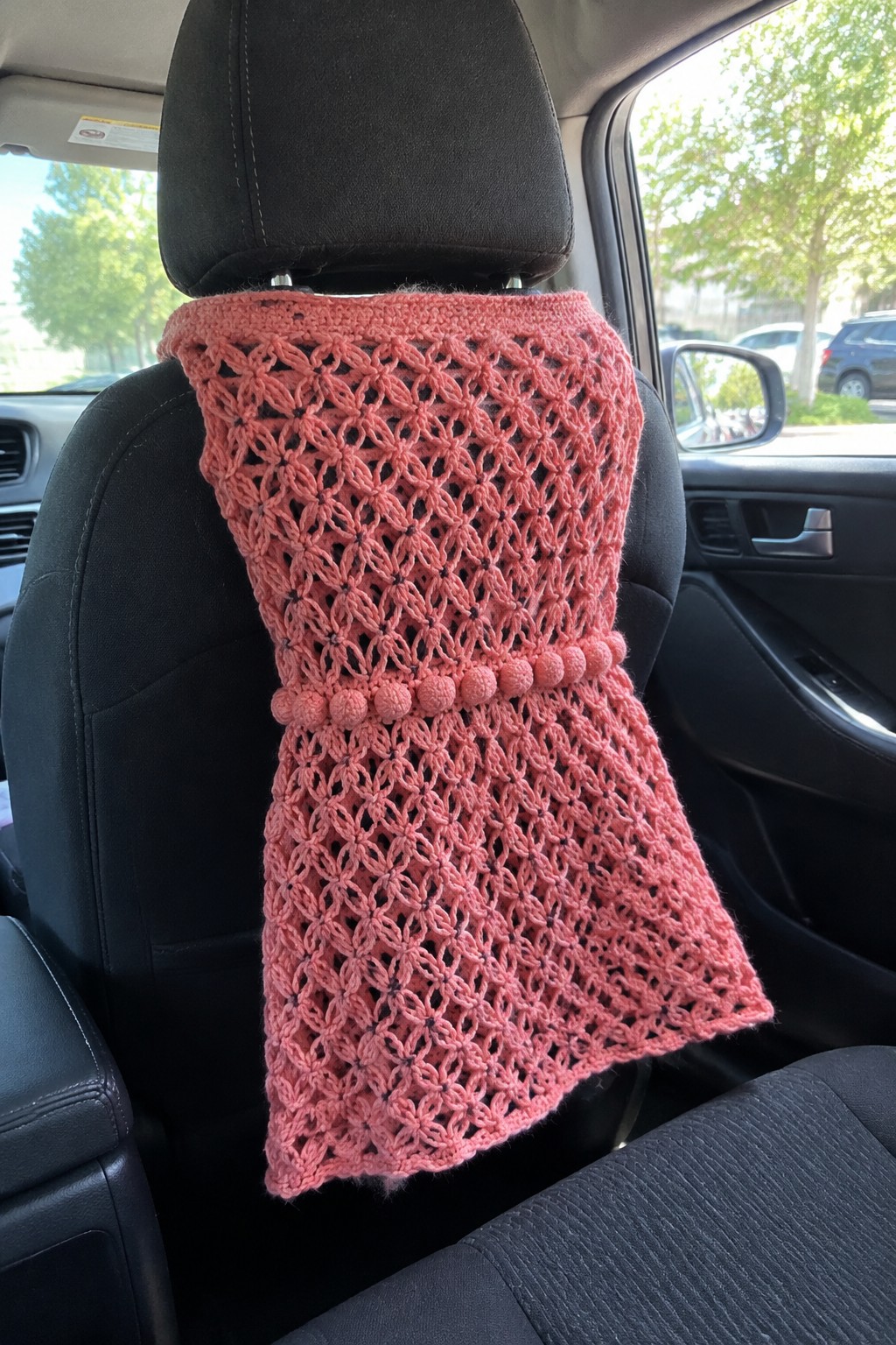 Pink crocheted lace cover with pom-poms on black car seat headrest