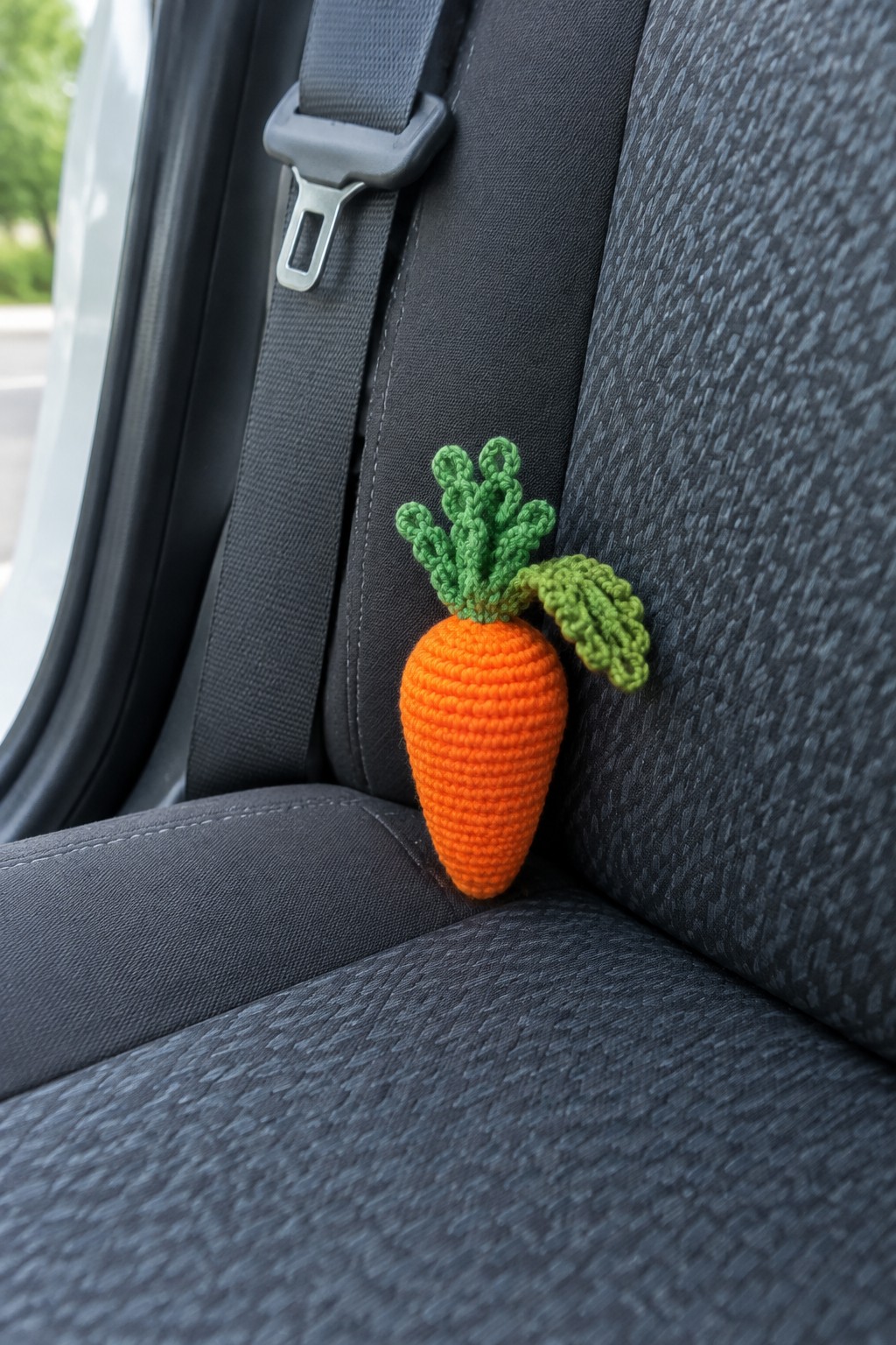 Orange crocheted carrot with green leaves on dark gray car seat beside seatbelt.