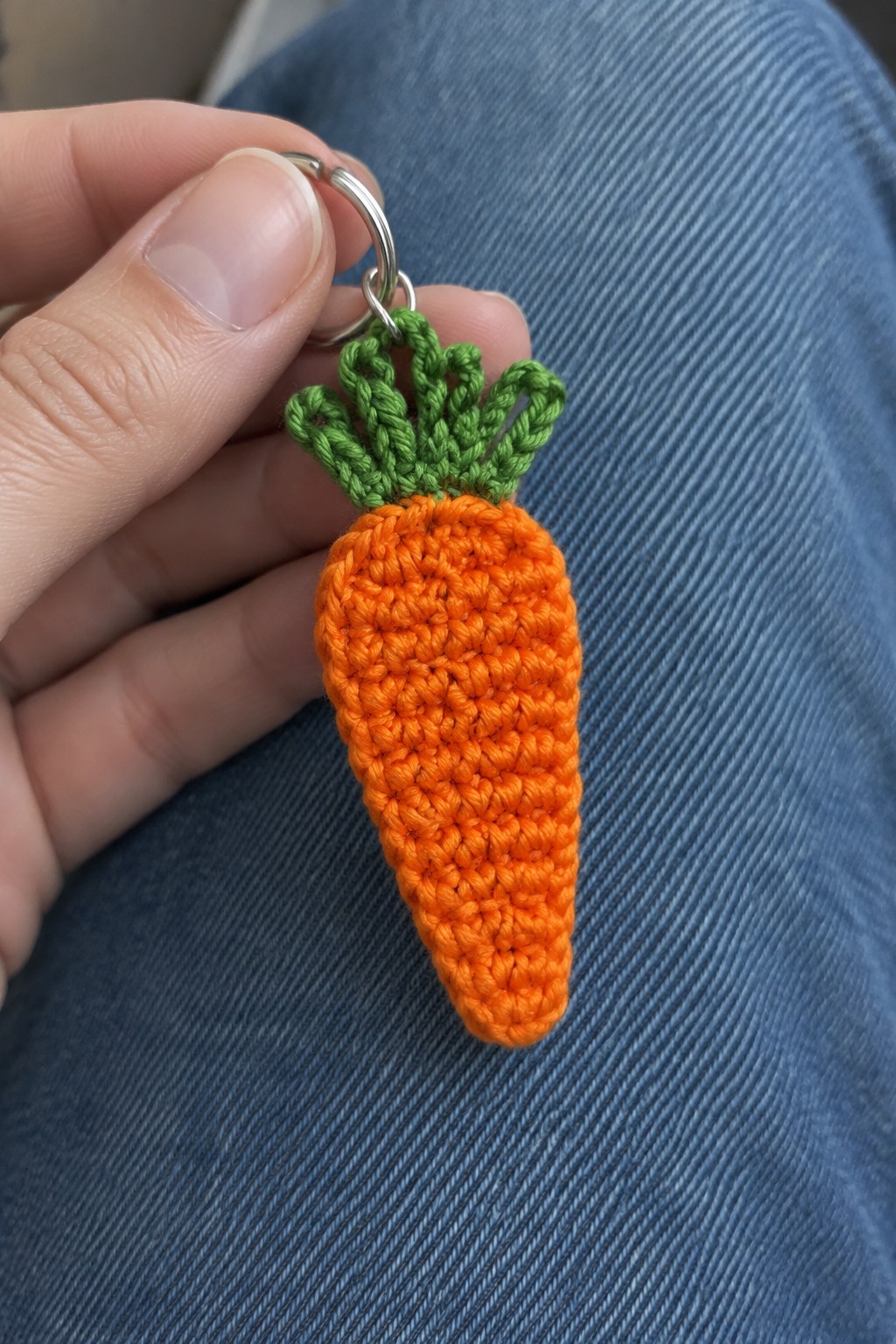 Hand holding crocheted orange carrot keychain with green top on blue denim