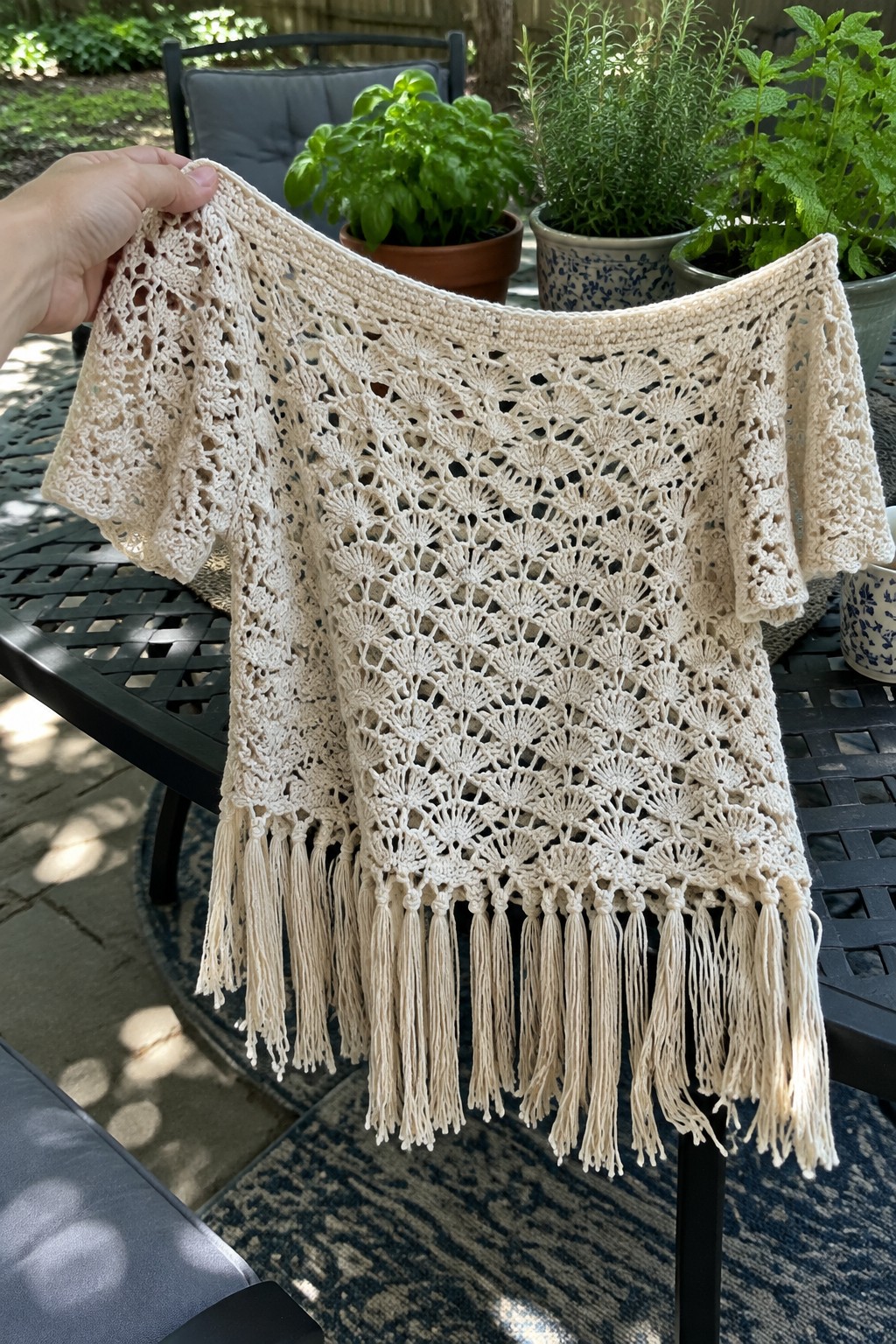 A cream crocheted top with fan lace patterns, short sleeves, and fringe hem held up outdoors over a black table.