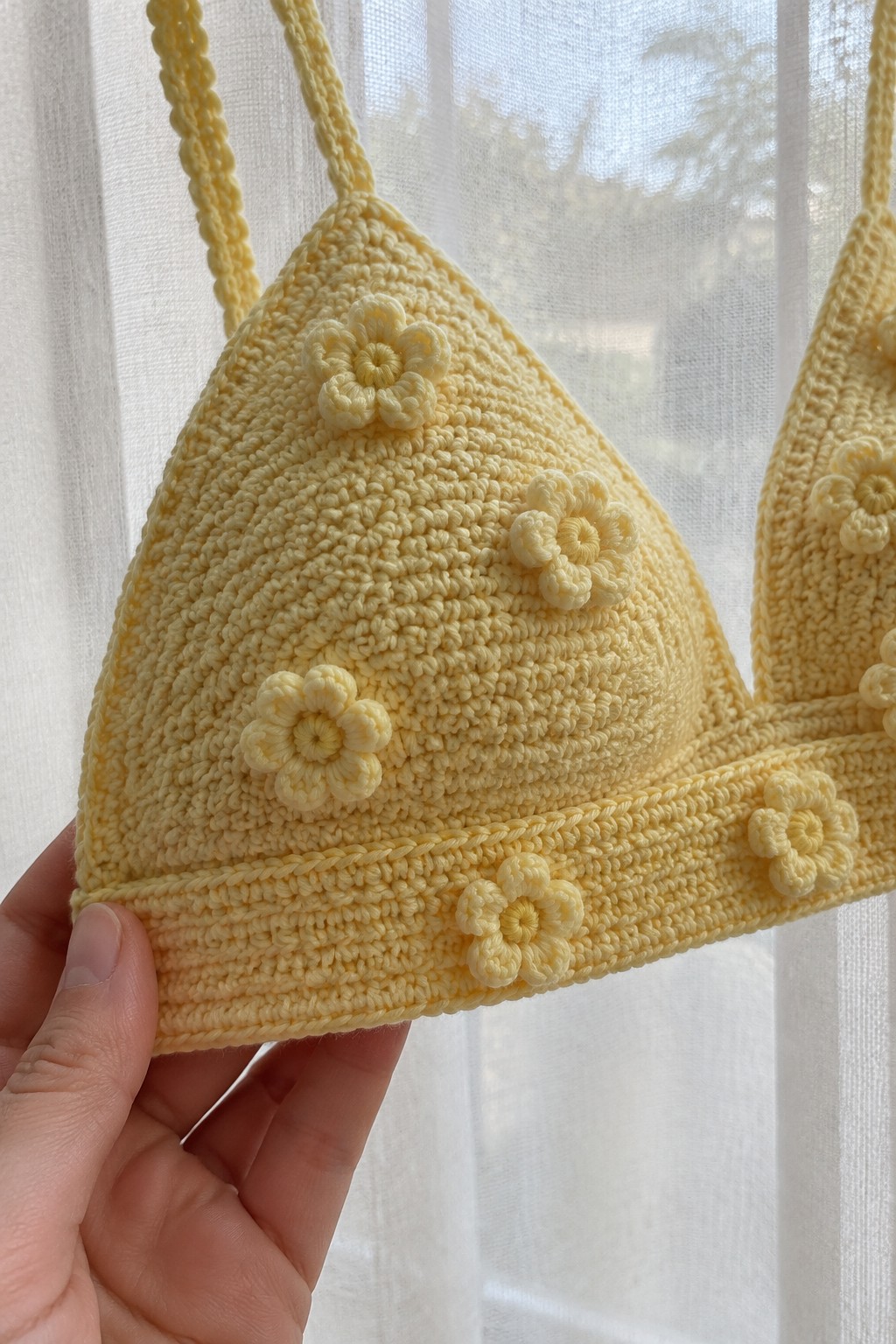 Hand holding pale yellow crocheted bikini top with flower appliqués by sheer curtain.
