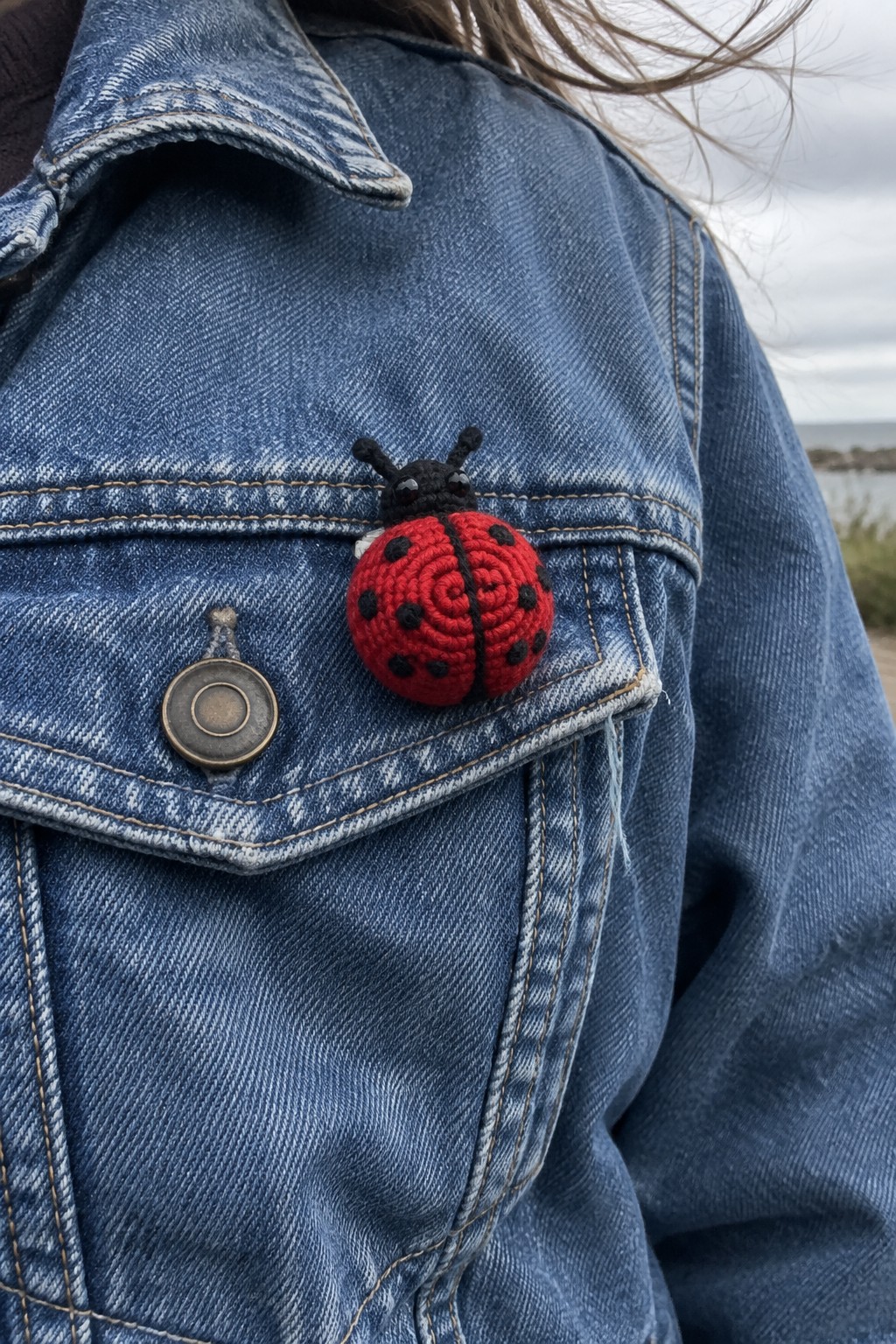 Red crocheted ladybug pin on blue denim jacket near pocket by water
