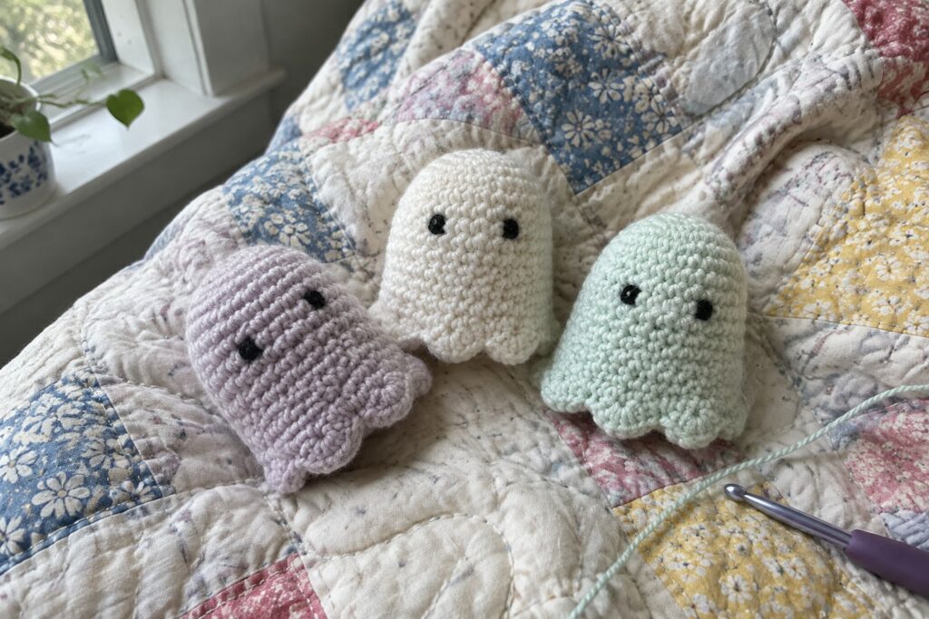 18 Playful Crochet Halloween Projects Built Around Ghosts, Pumpkins, and Bats Three pastel crocheted ghosts on floral quilt with notebook, hook, and yarn.