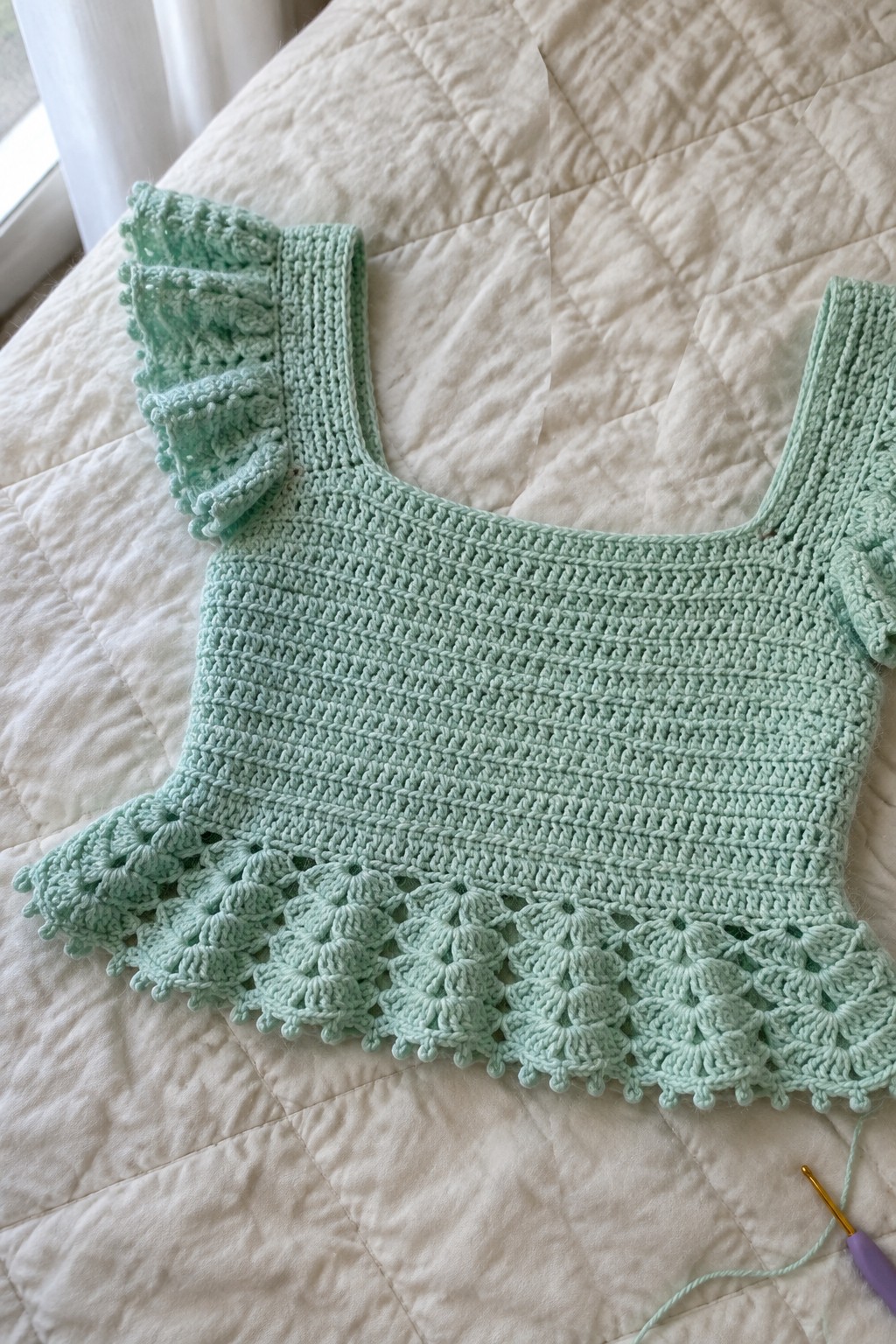 Mint green crocheted crop top with ruffled shoulders and scalloped hem on white quilt