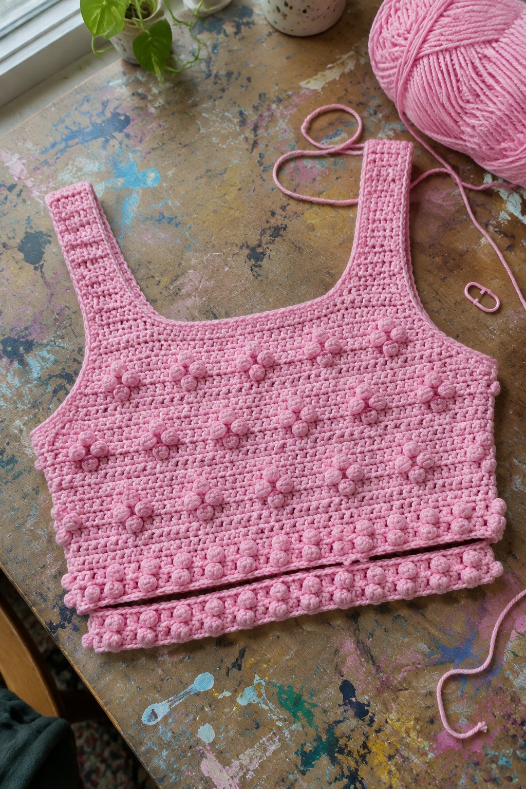 Pink crocheted tank top with bobble texture on paint-splattered table beside pink yarn.