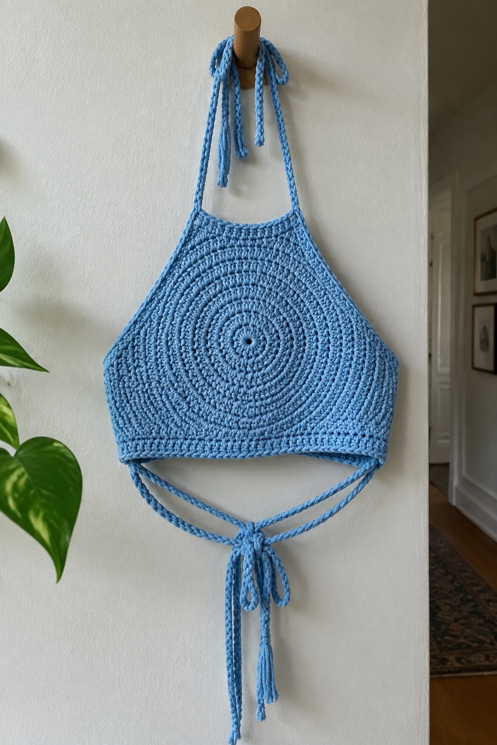 A light blue crocheted halter crop top with a circular center pattern hangs from a wooden peg, showing long neck ties and waist ties with tassels.