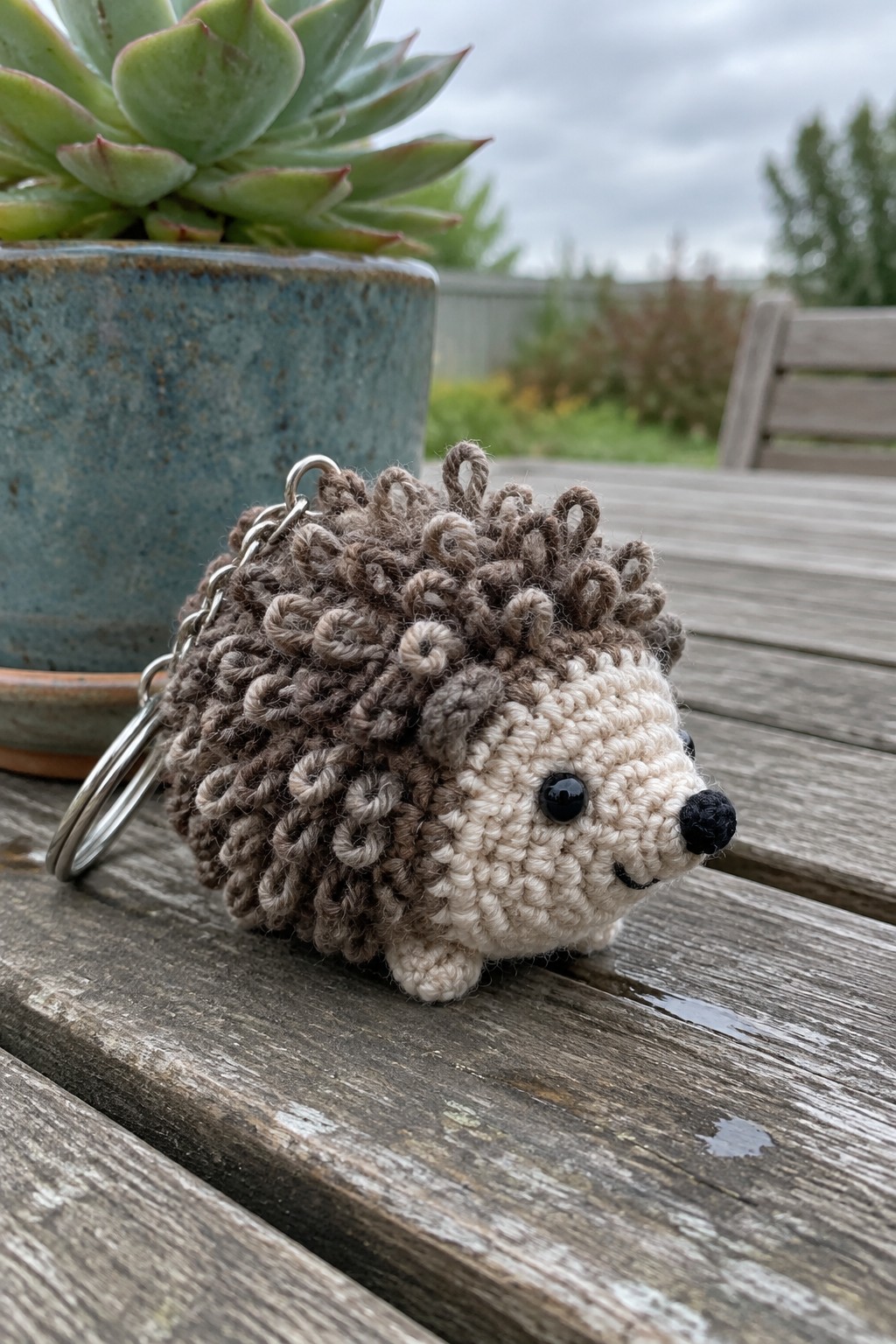 Crocheted brown and beige hedgehog keychain on wet wooden table beside succulent pot