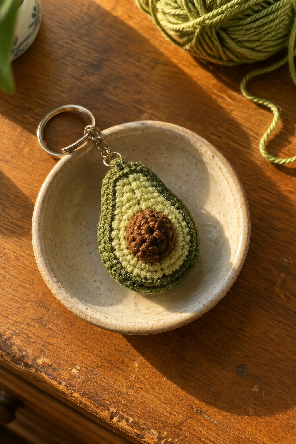 Crocheted avocado keychain resting in a ceramic bowl on a wooden table with green yarn in the background.