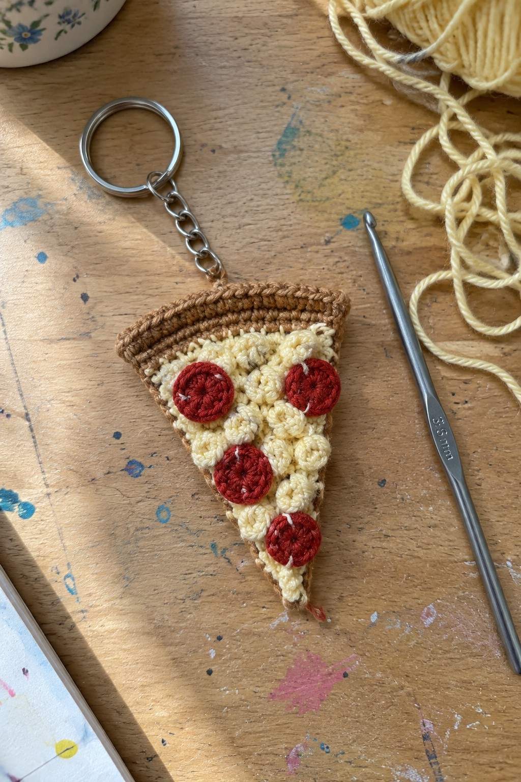 Crocheted pizza slice keychain with red pepperoni beside silver hook and yellow yarn