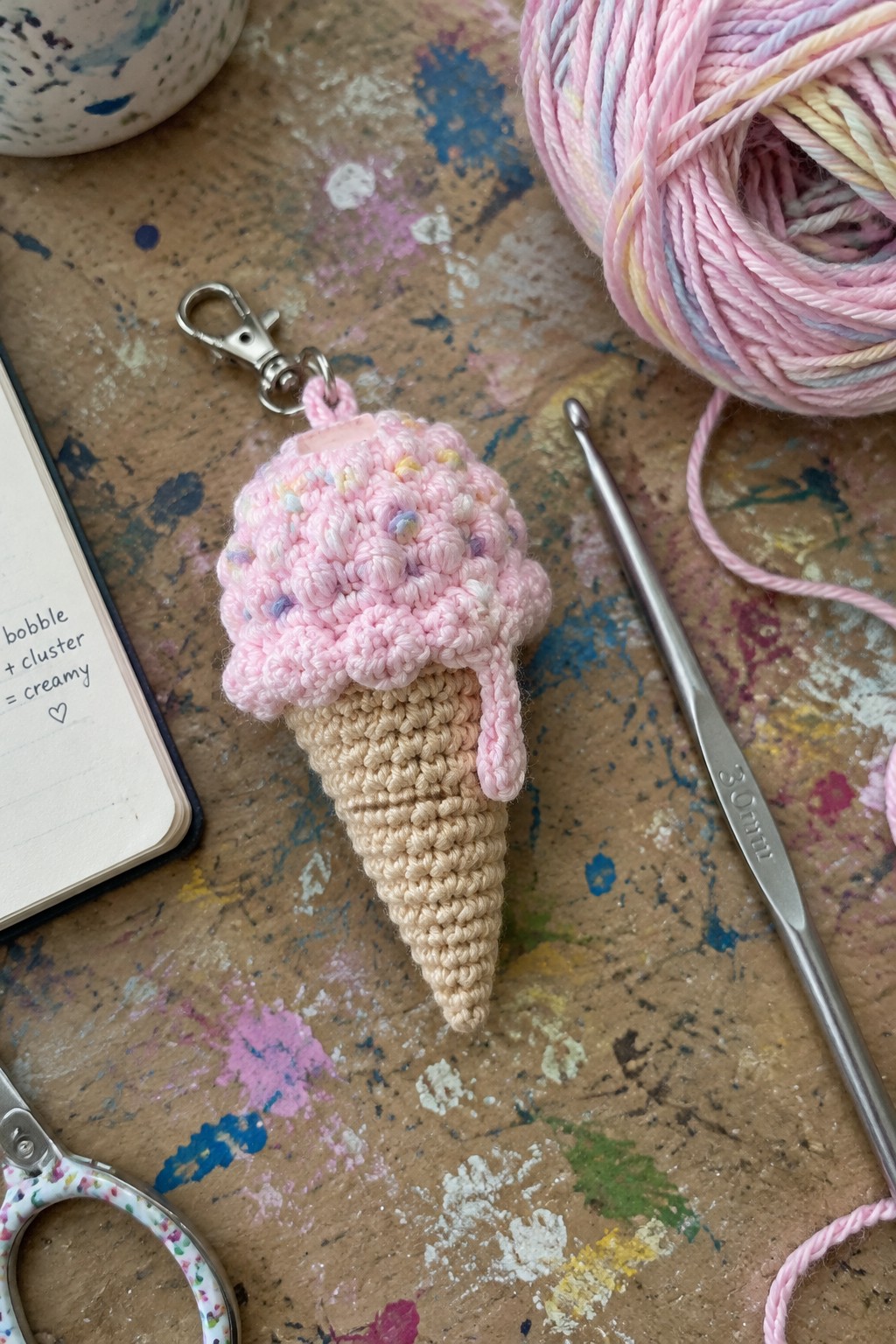 Crocheted pink ice cream cone keychain beside yarn, hook, and notebook on painted table