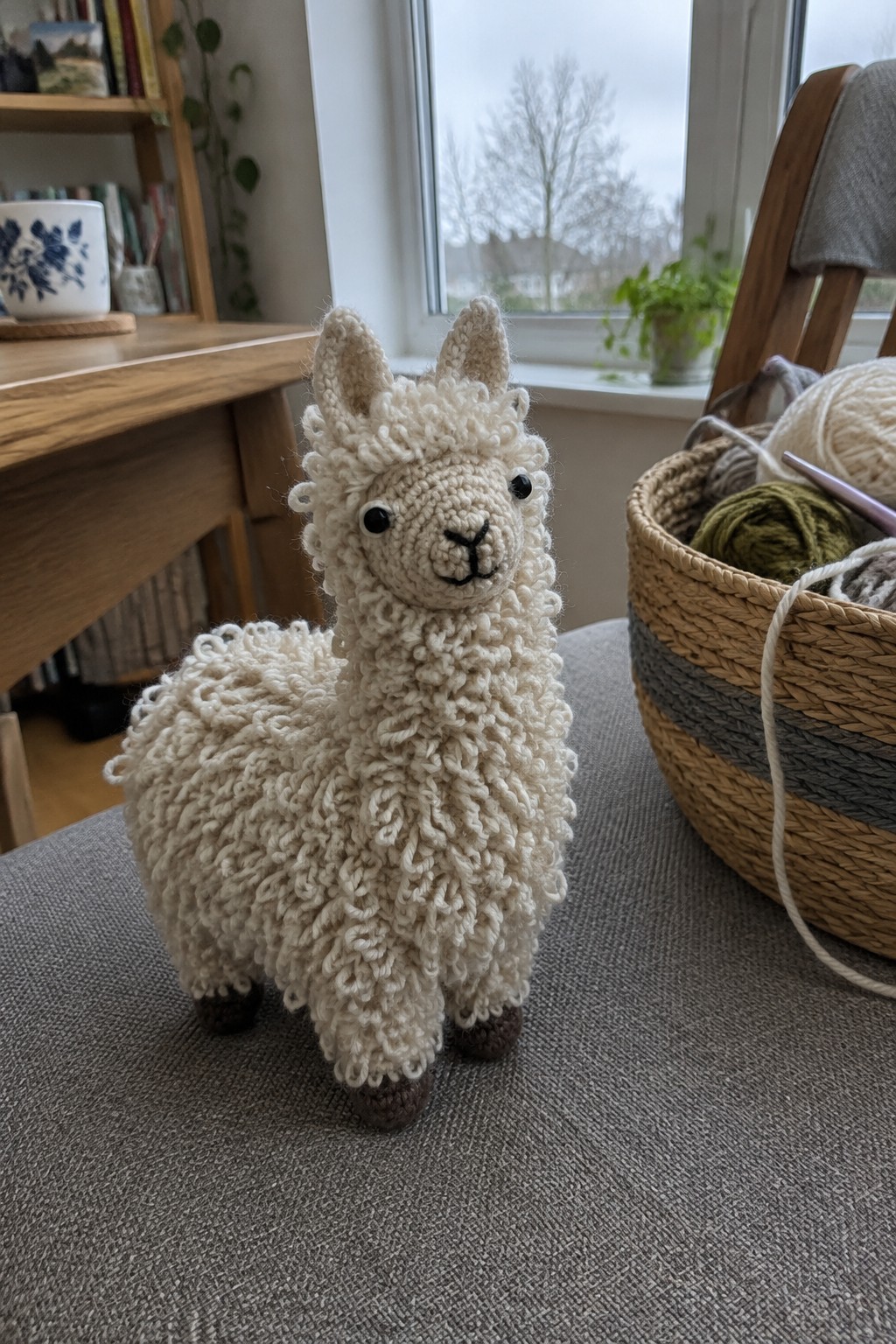 Crocheted white llama beside basket of yarn on gray fabric chair