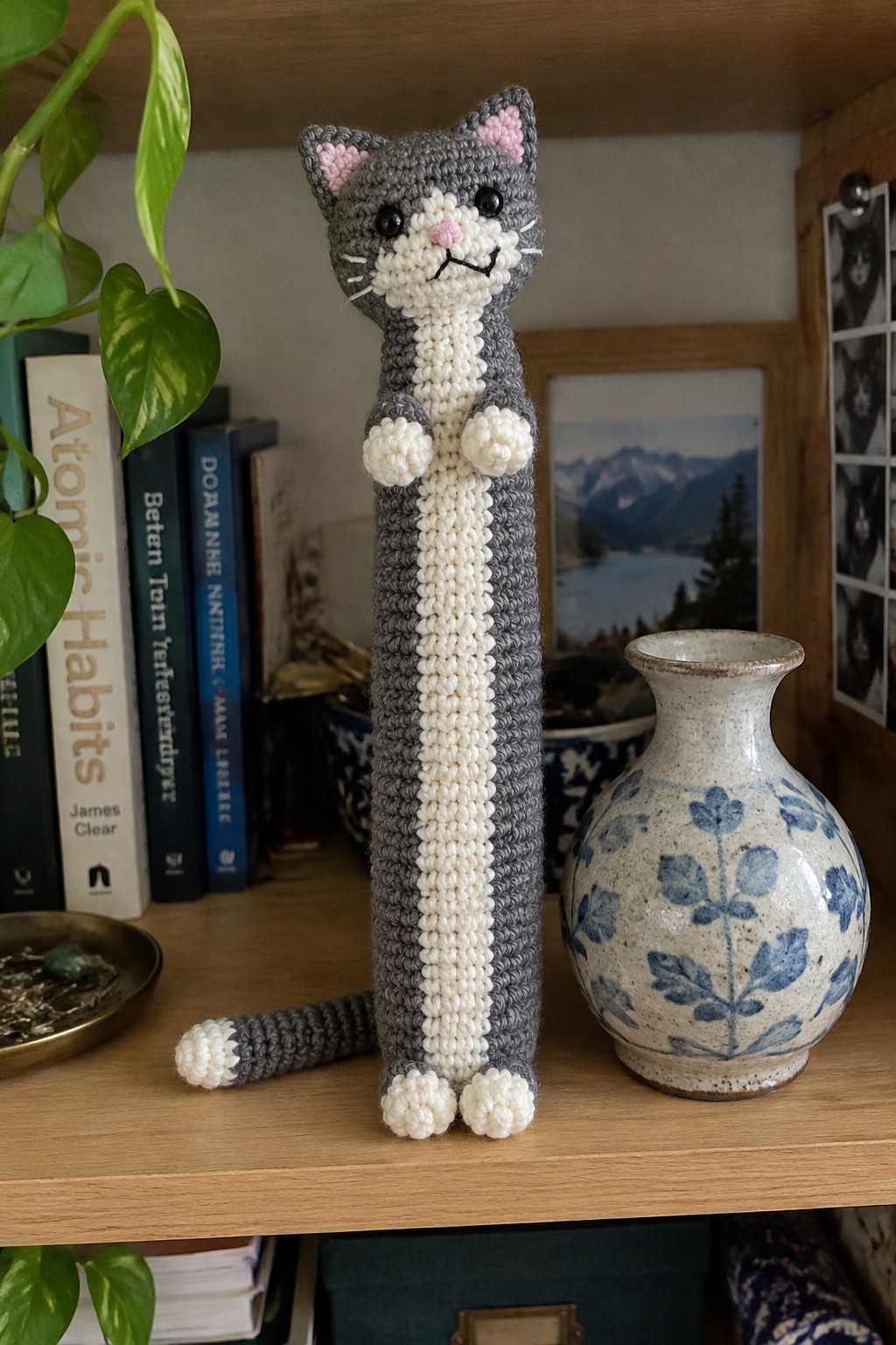Crocheted gray and white cat on wooden shelf beside floral vase and books