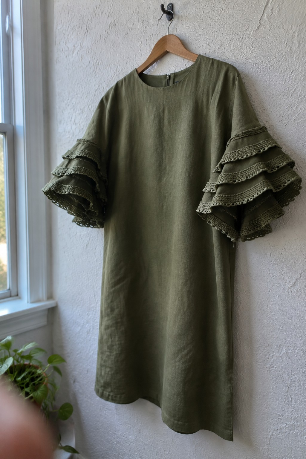 Olive green linen dress with tiered lace sleeves on wooden hanger against white wall