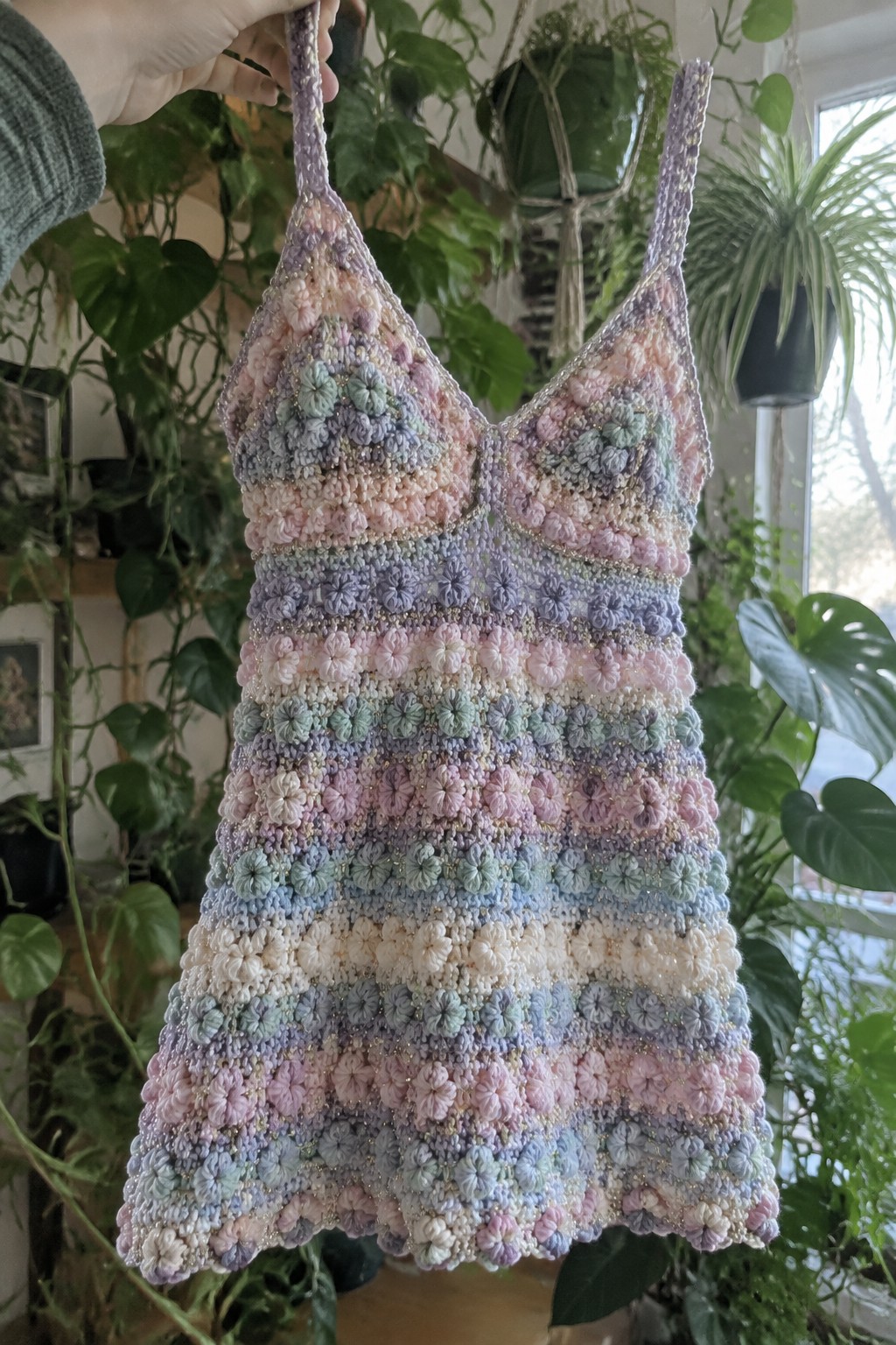 Hand holding pastel crocheted floral dress with beads amid lush green plants