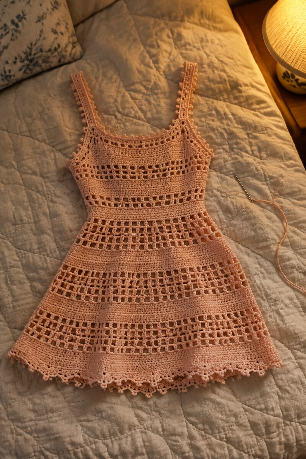 Peach crocheted dress with openwork pattern on quilted bed beside needle