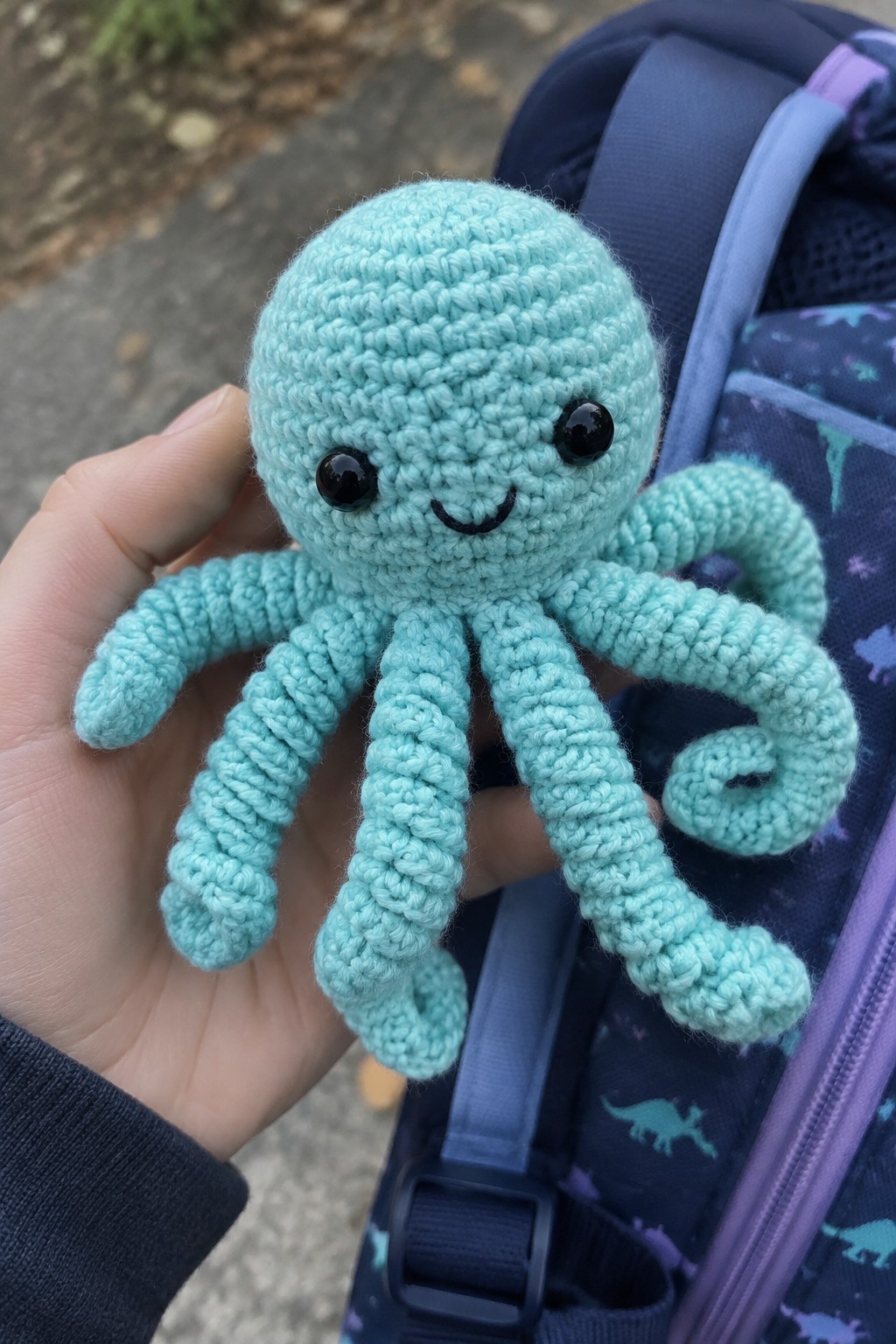 Hand holding light blue crocheted octopus beside dinosaur-patterned blue backpack
