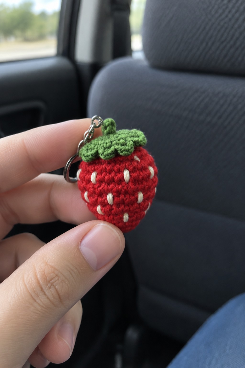 Hand holding crocheted red strawberry keychain with white seeds and green leaves in car