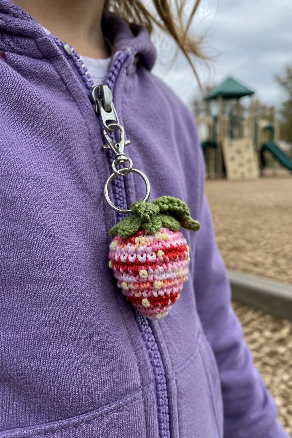 Purple hoodie with pink crocheted strawberry keychain clipped to zipper at playground