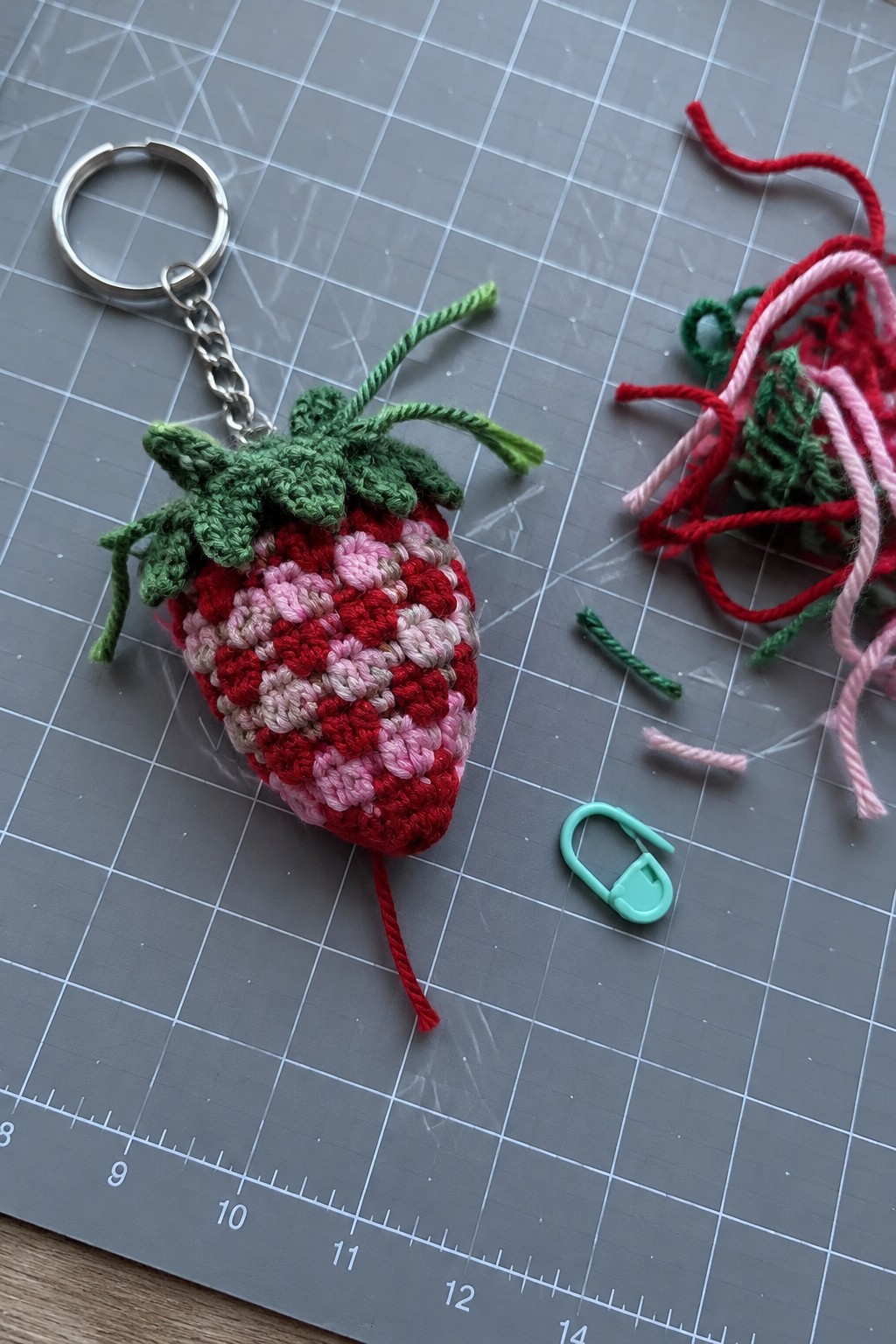Crocheted red-pink strawberry keychain with green top on gray gridded mat with yarn scraps