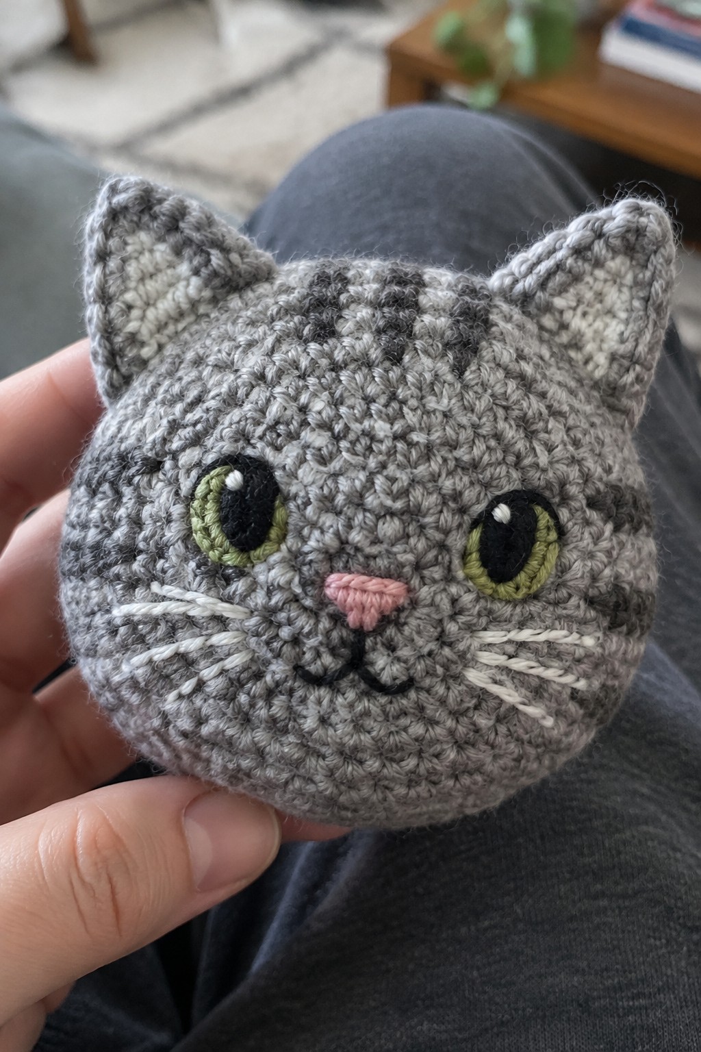 Hand holding crocheted gray tabby cat head with green eyes and pink nose