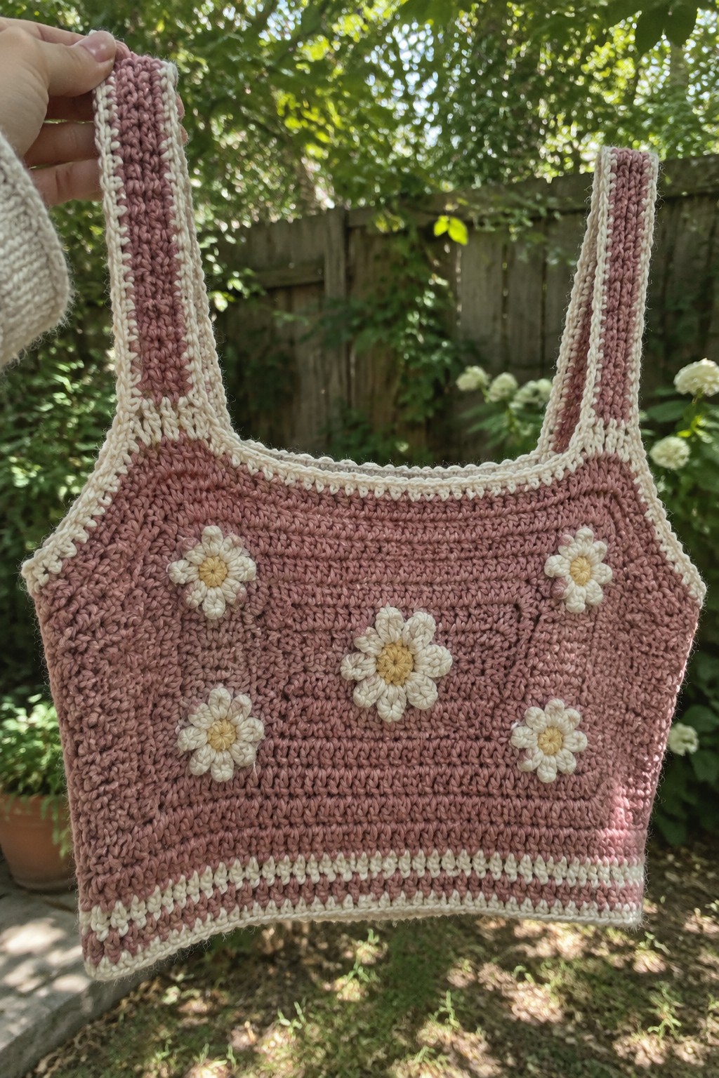 Hand holding pink crocheted crop top with daisy flowers and white trim outdoors