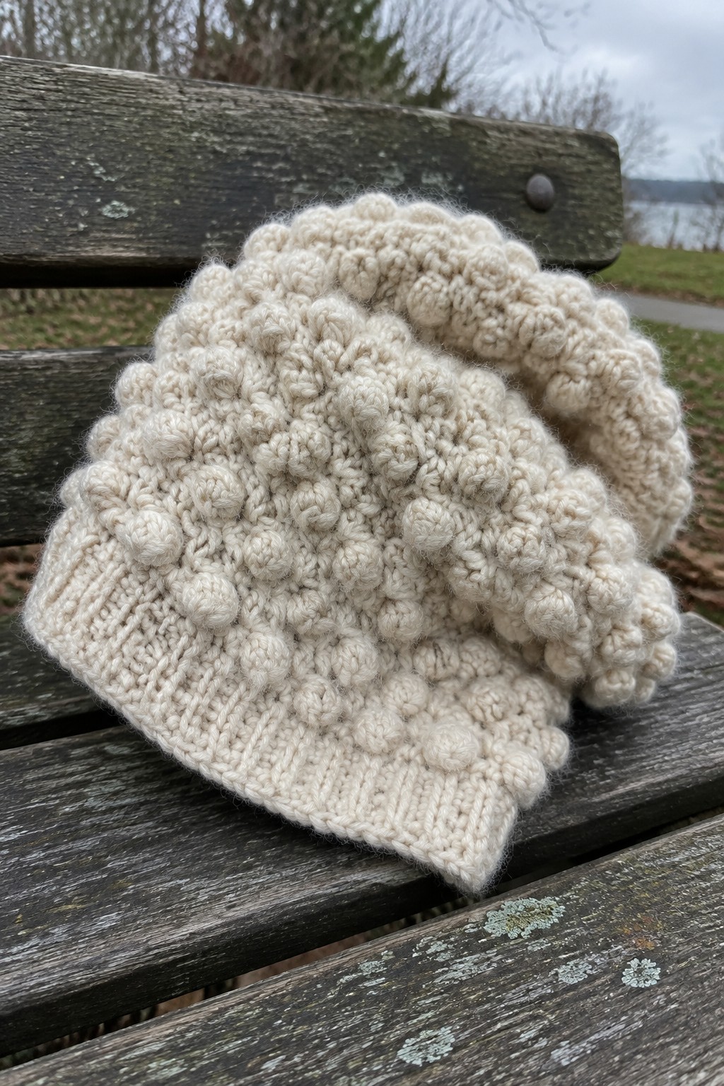 Cream bobble-knit beanie resting on weathered wooden bench outdoors.