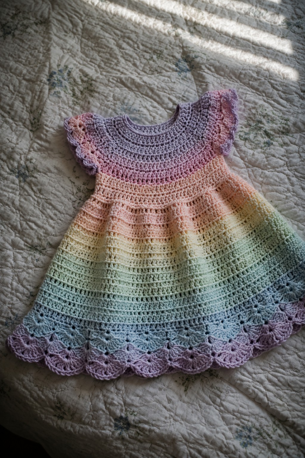Rainbow crocheted child's dress with ruffles on quilted bed in sunlight.