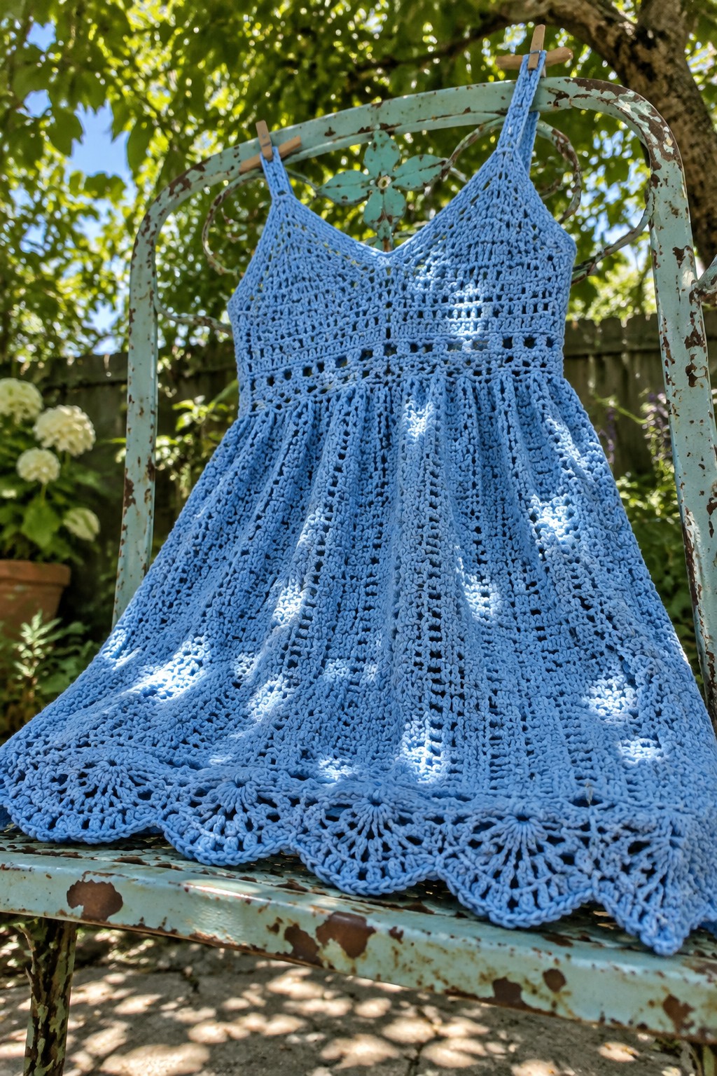 A light blue crocheted child's dress with thin straps and a scalloped hem hangs on a weathered metal chair outdoors.