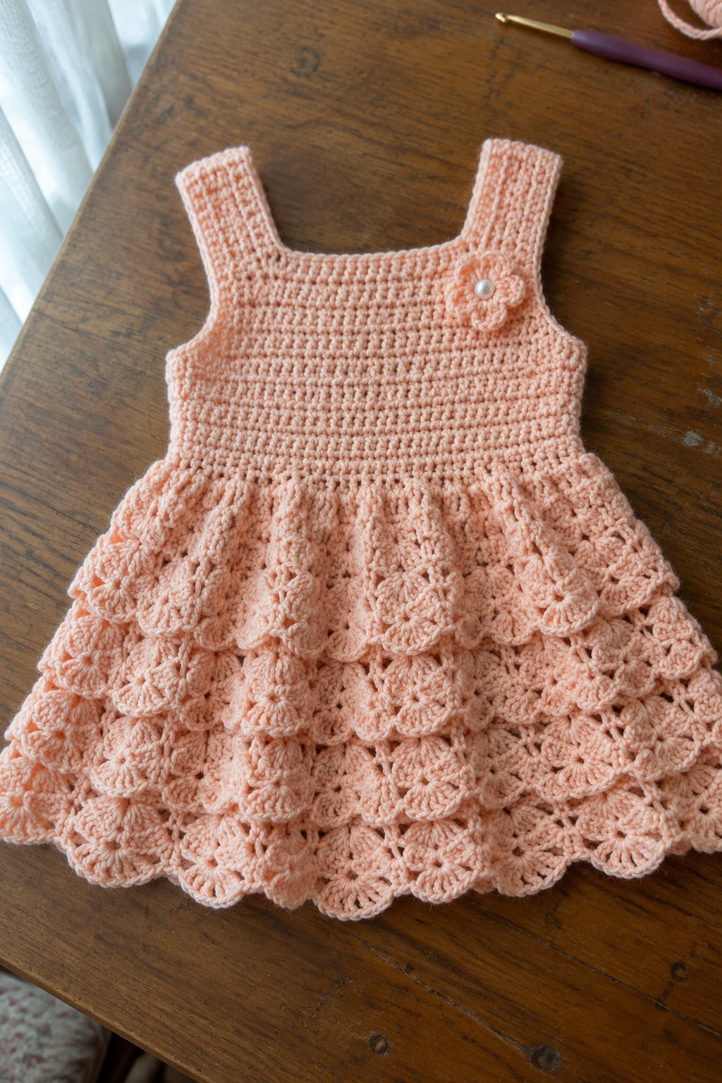 Peach crocheted toddler dress with flower detail on wooden table beside hook and yarn