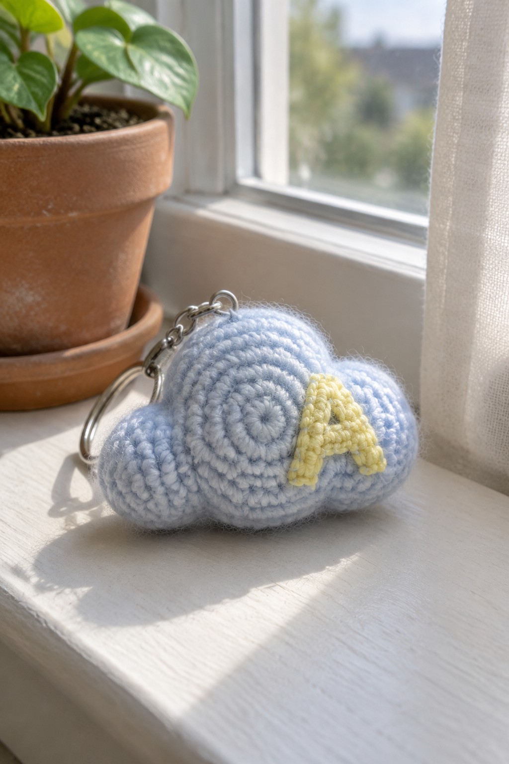 Light blue crocheted cloud keychain with yellow A on white windowsill