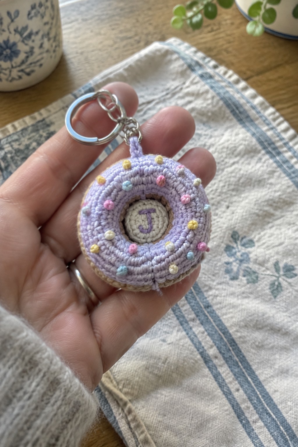 Hand holds crocheted lavender donut keychain with pastel sprinkles and letter J