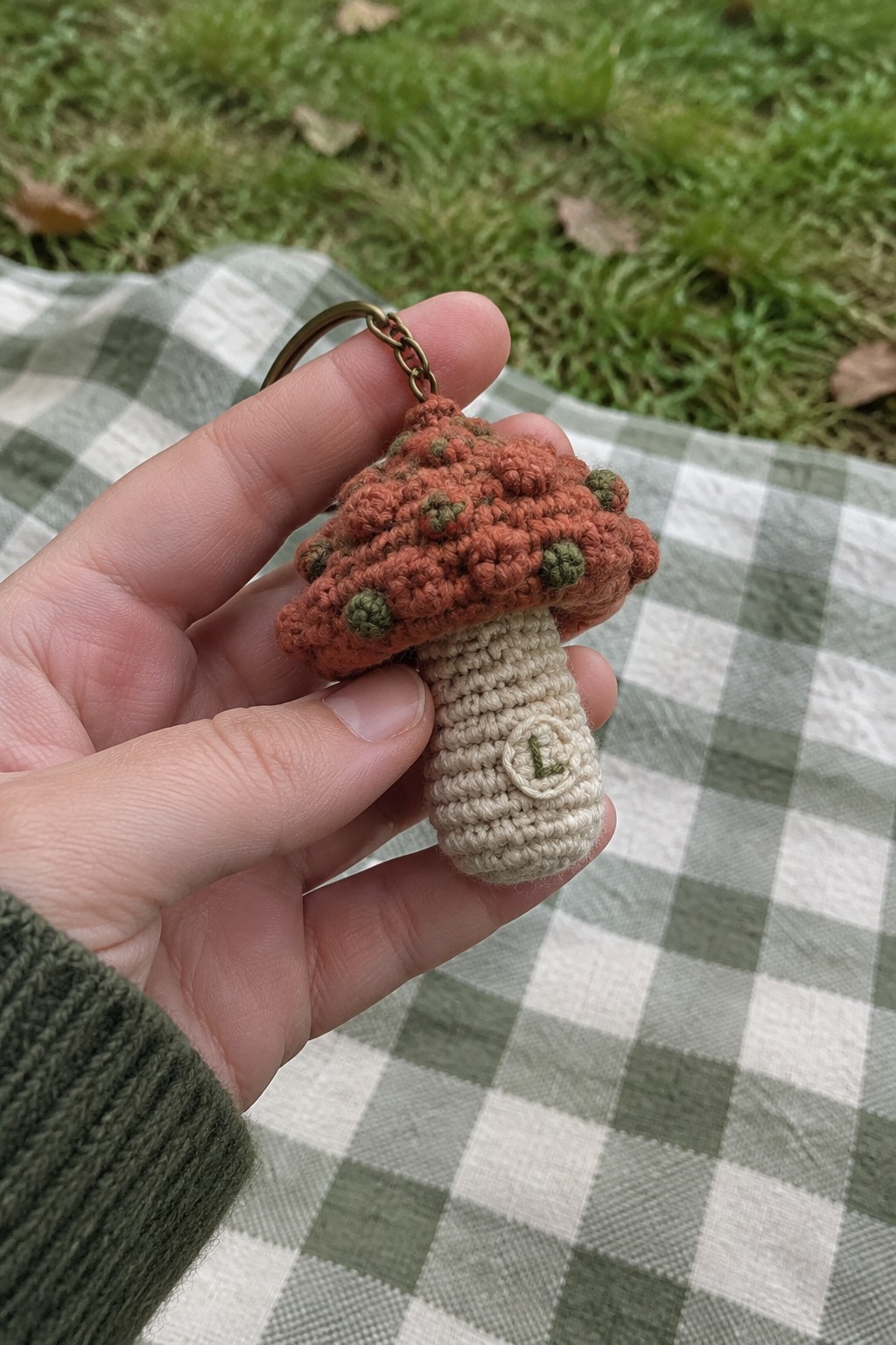 Hand holding crocheted mushroom keychain with rust cap and green spots on checkered blanket