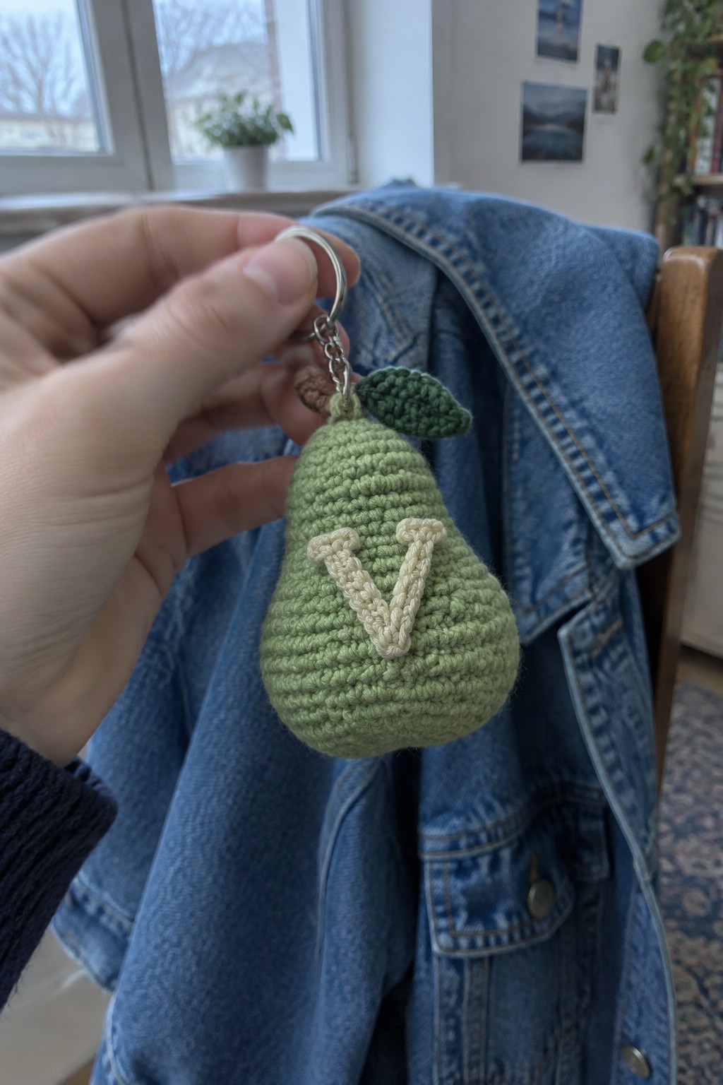 Hand holding light green crocheted pear keychain with beige V on denim jacket