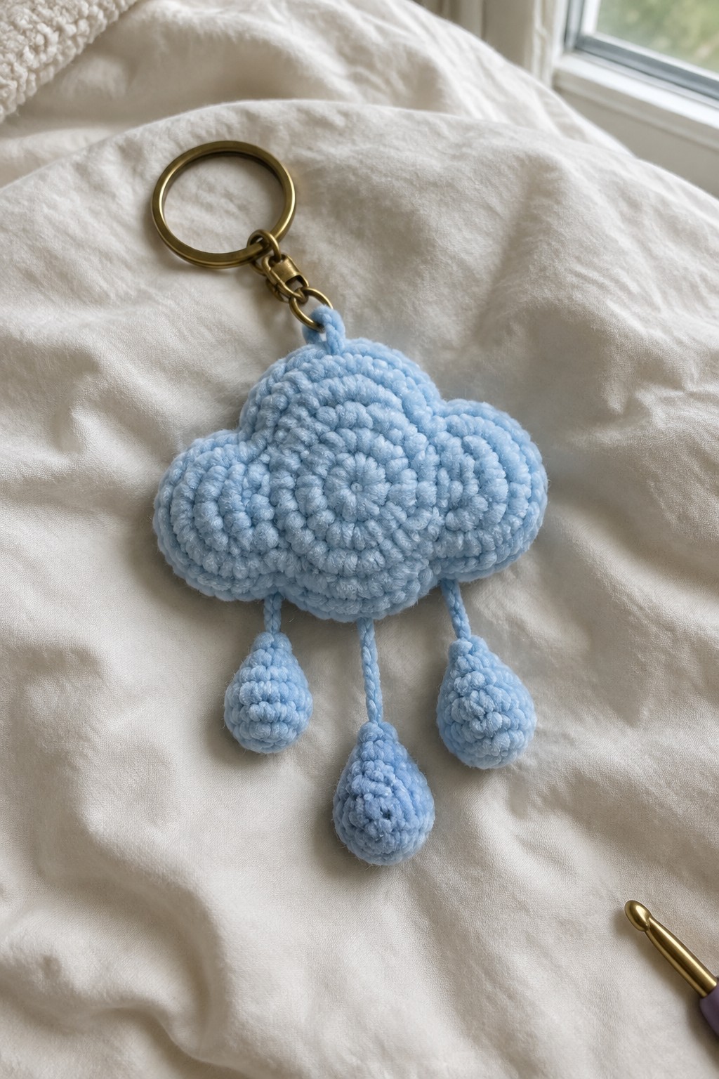Light blue crocheted cloud keychain with three dangling raindrops on white fabric