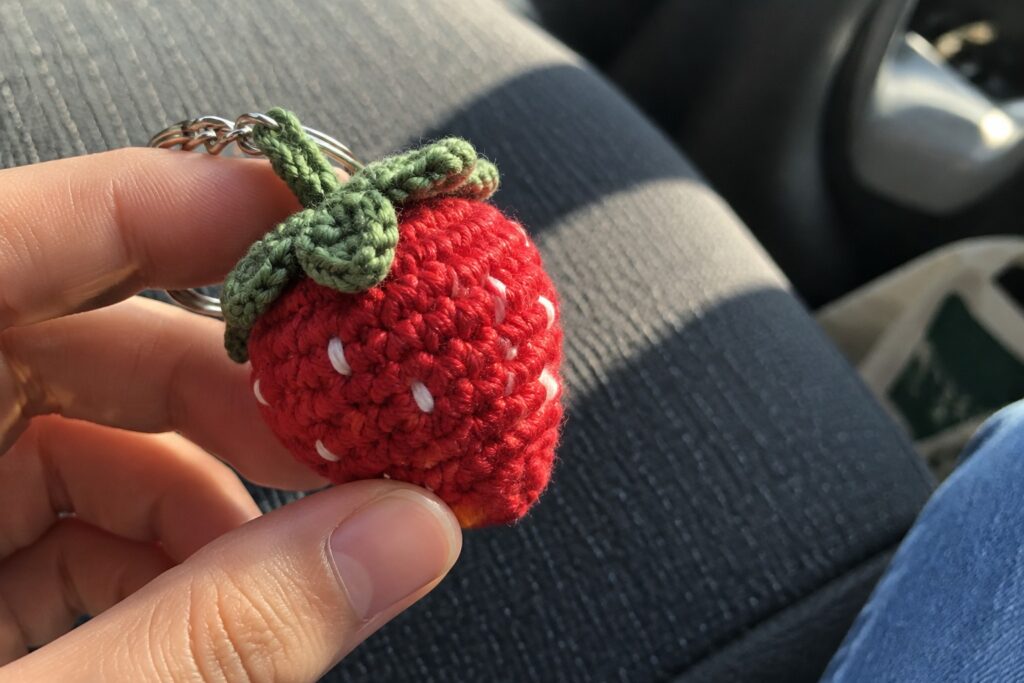 Hand holding crocheted red strawberry keychain with green leaves inside a car.