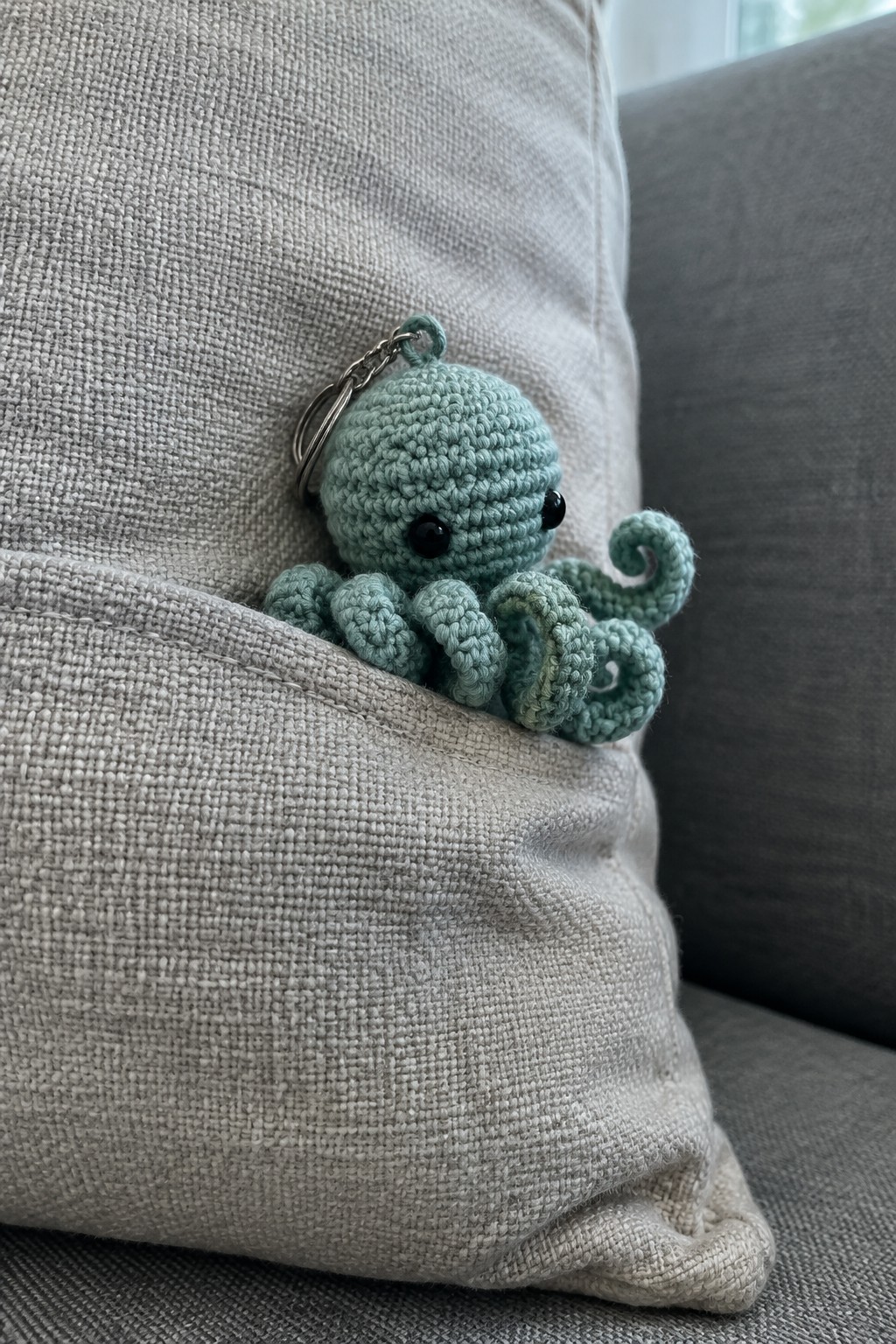 Mint green crocheted octopus keychain with black eyes peeking from beige pillow pocket