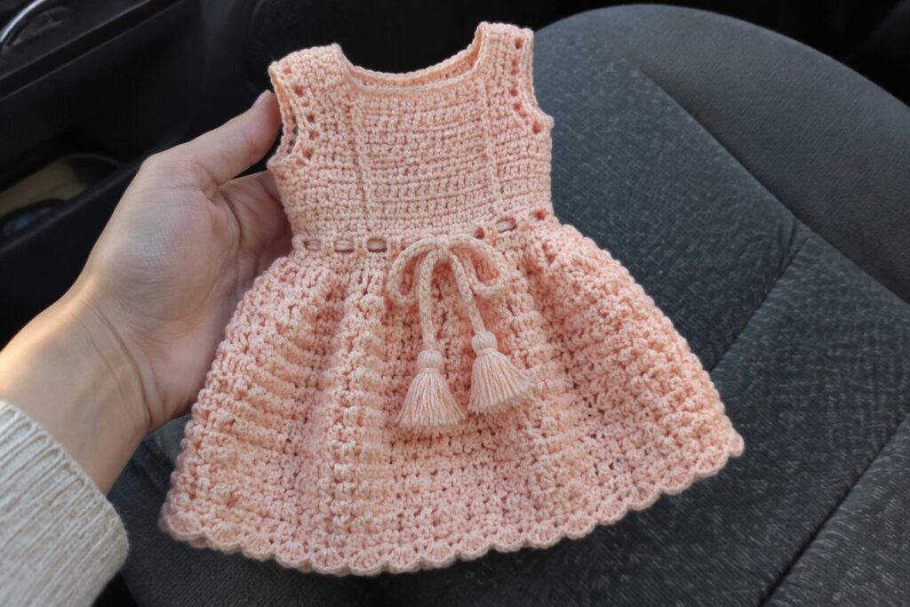 20 Crochet Dresses With Fitted Bodices and Adjustable Ties Hand holding peach crocheted dress with bow and tassels over car seat