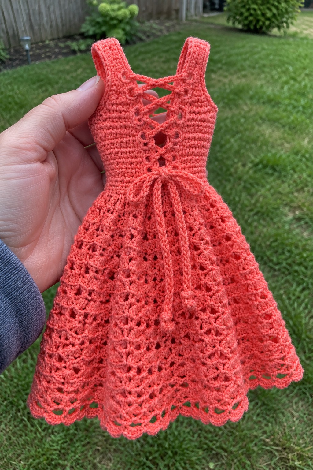 Hand holding tiny coral crocheted dress with lace-up back, bow, and lacy skirt.