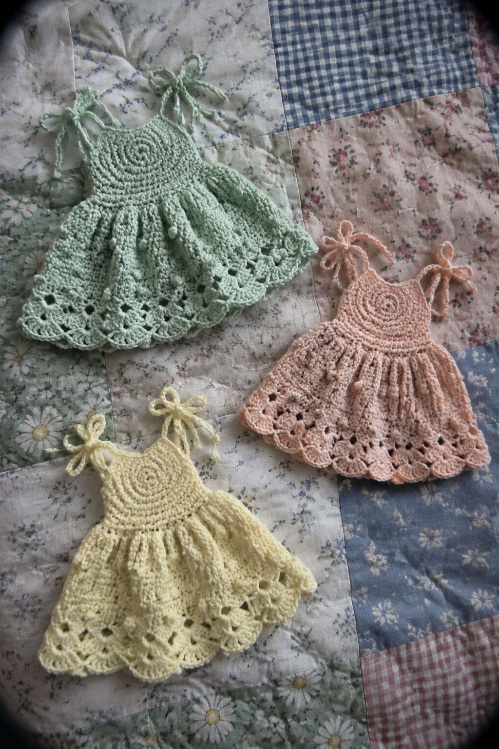 Three pastel crocheted baby dresses with bows on patchwork quilt