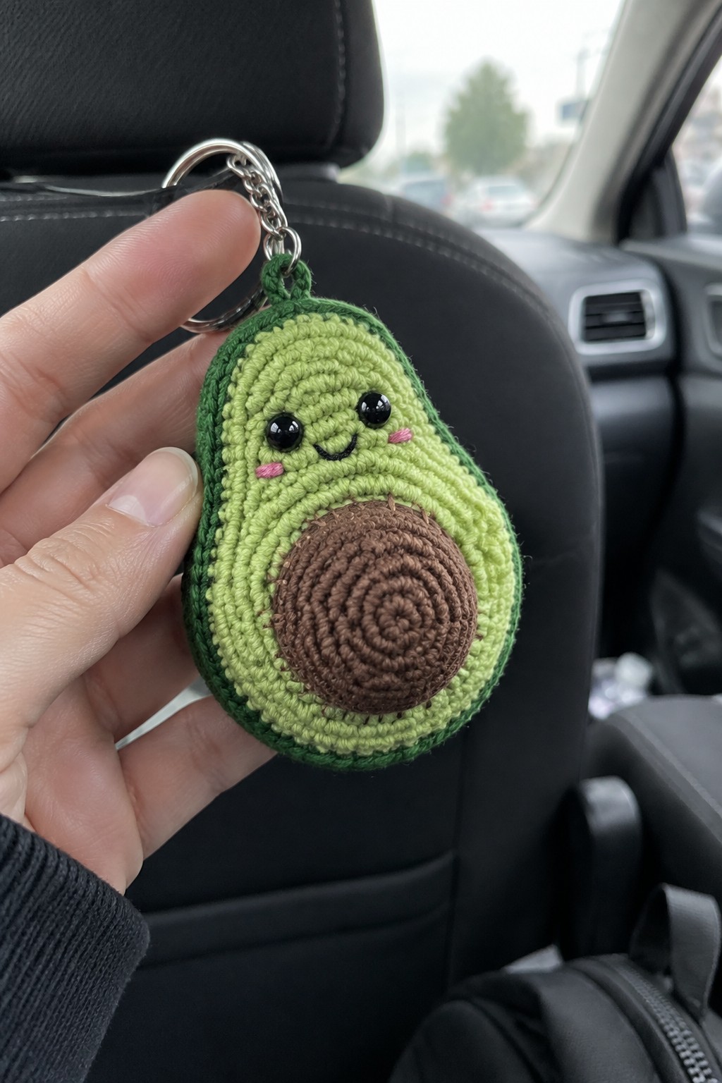 Hand holding smiling crocheted avocado keychain inside car interior