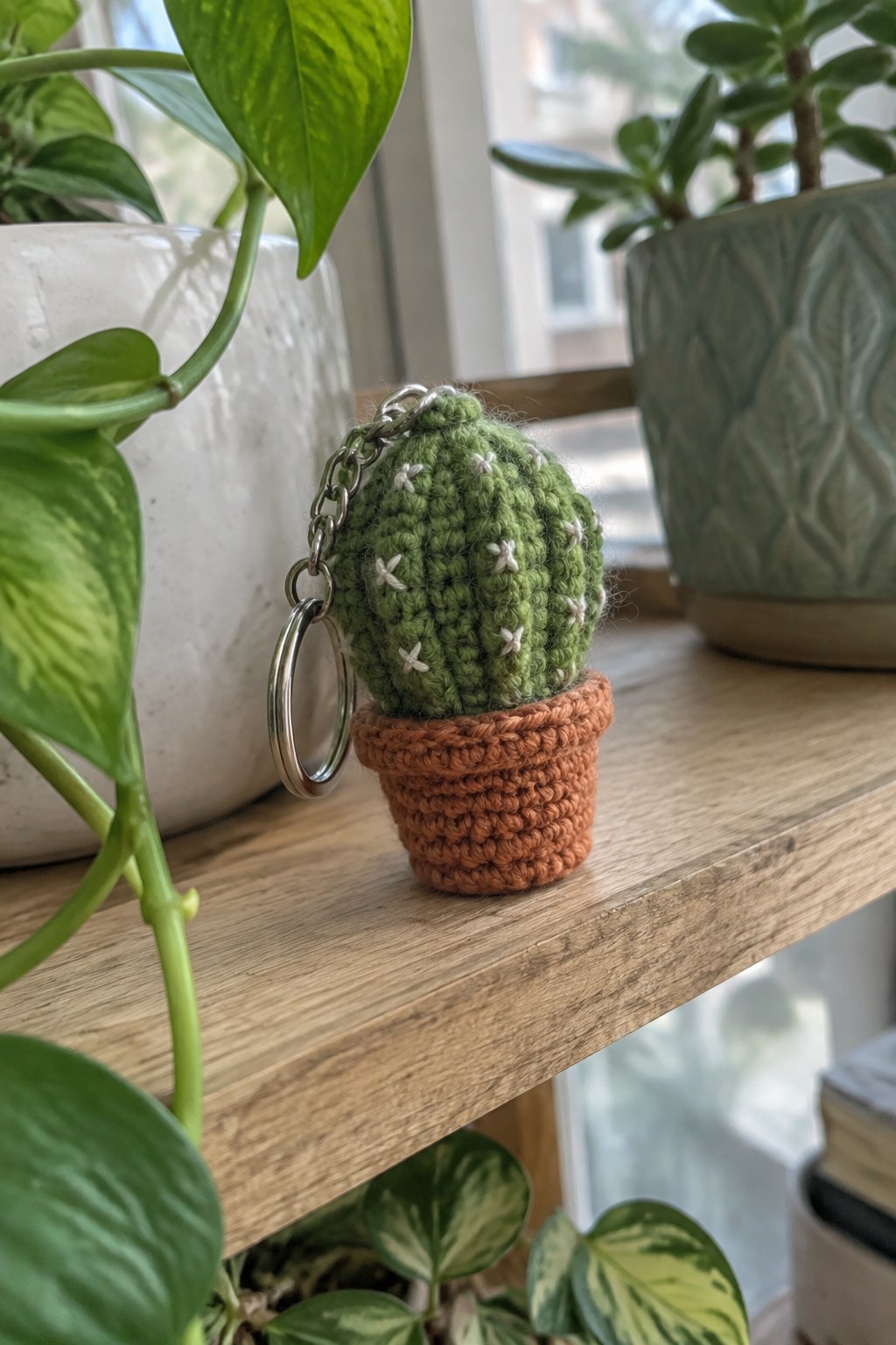 Crocheted green cactus keychain in orange pot on wooden shelf with plants