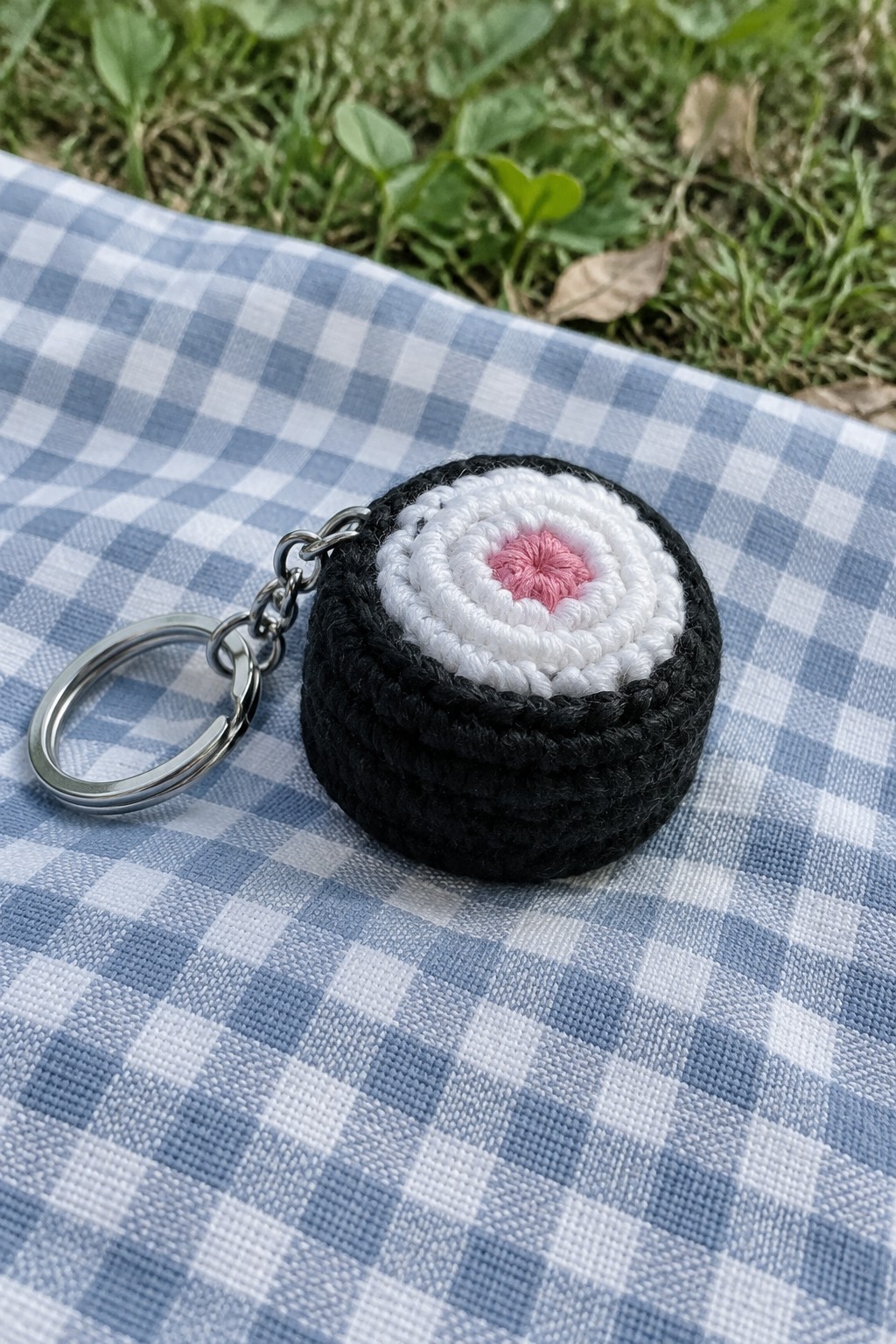 Crocheted sushi keychain with black nori, white rice, and pink center on checkered cloth.