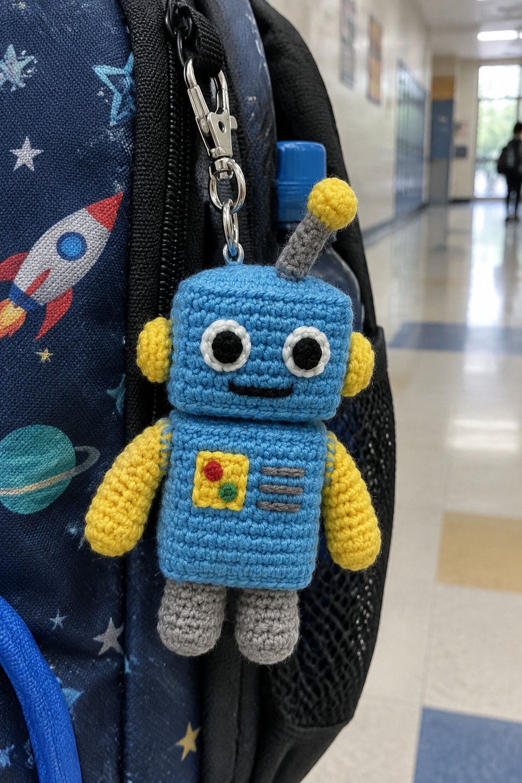 Blue crocheted robot keychain clipped to space-themed backpack in hallway