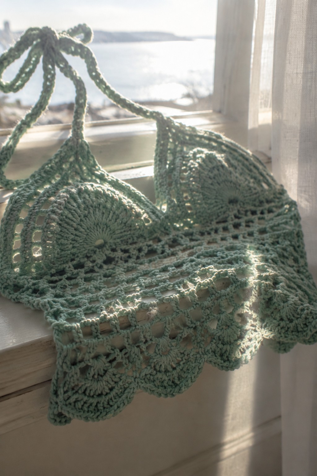 A light green crocheted halter top with circular lace motifs rests on a wooden windowsill.