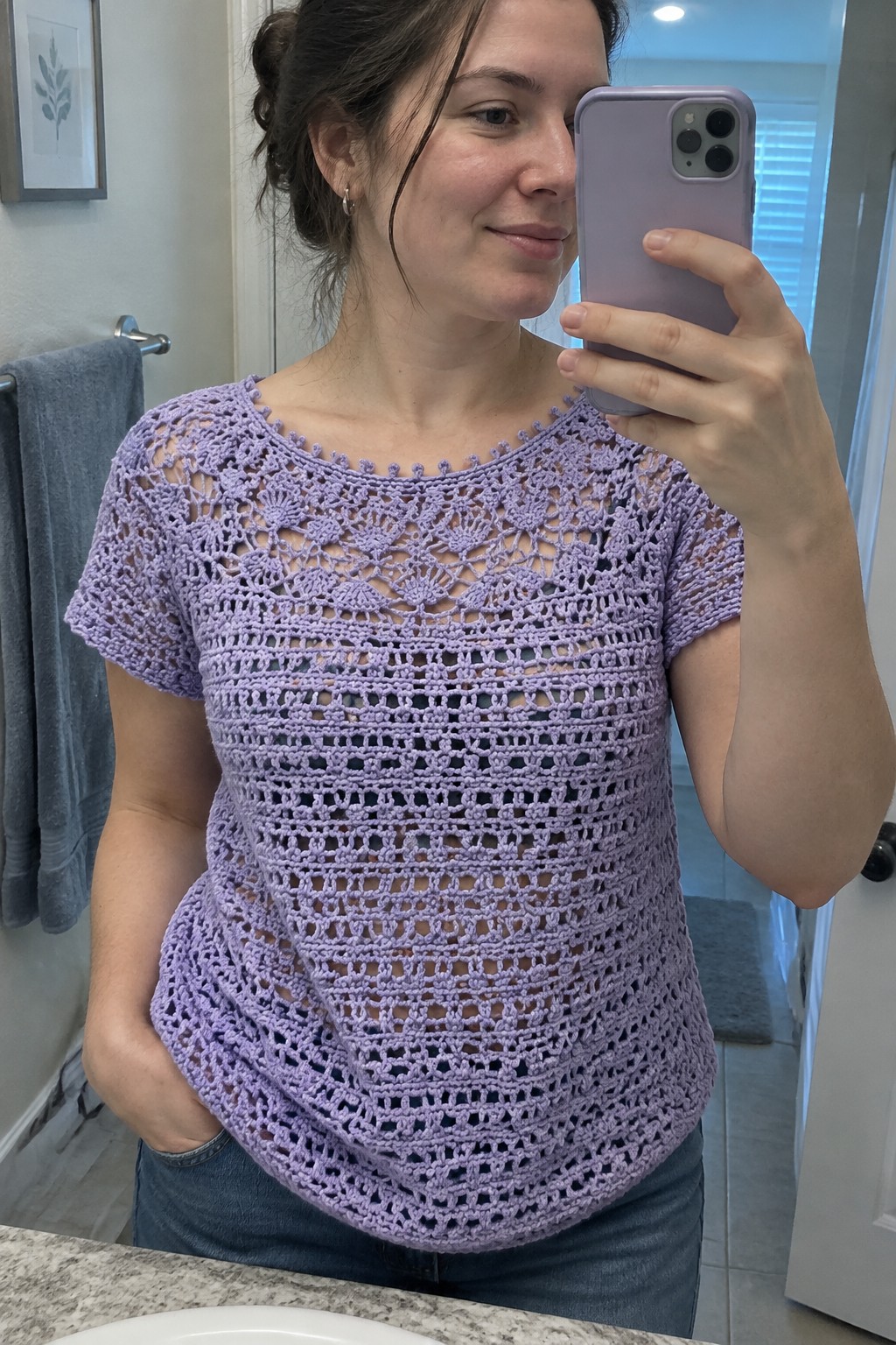 Woman in lavender open-crochet top taking bathroom mirror selfie with purple phone
